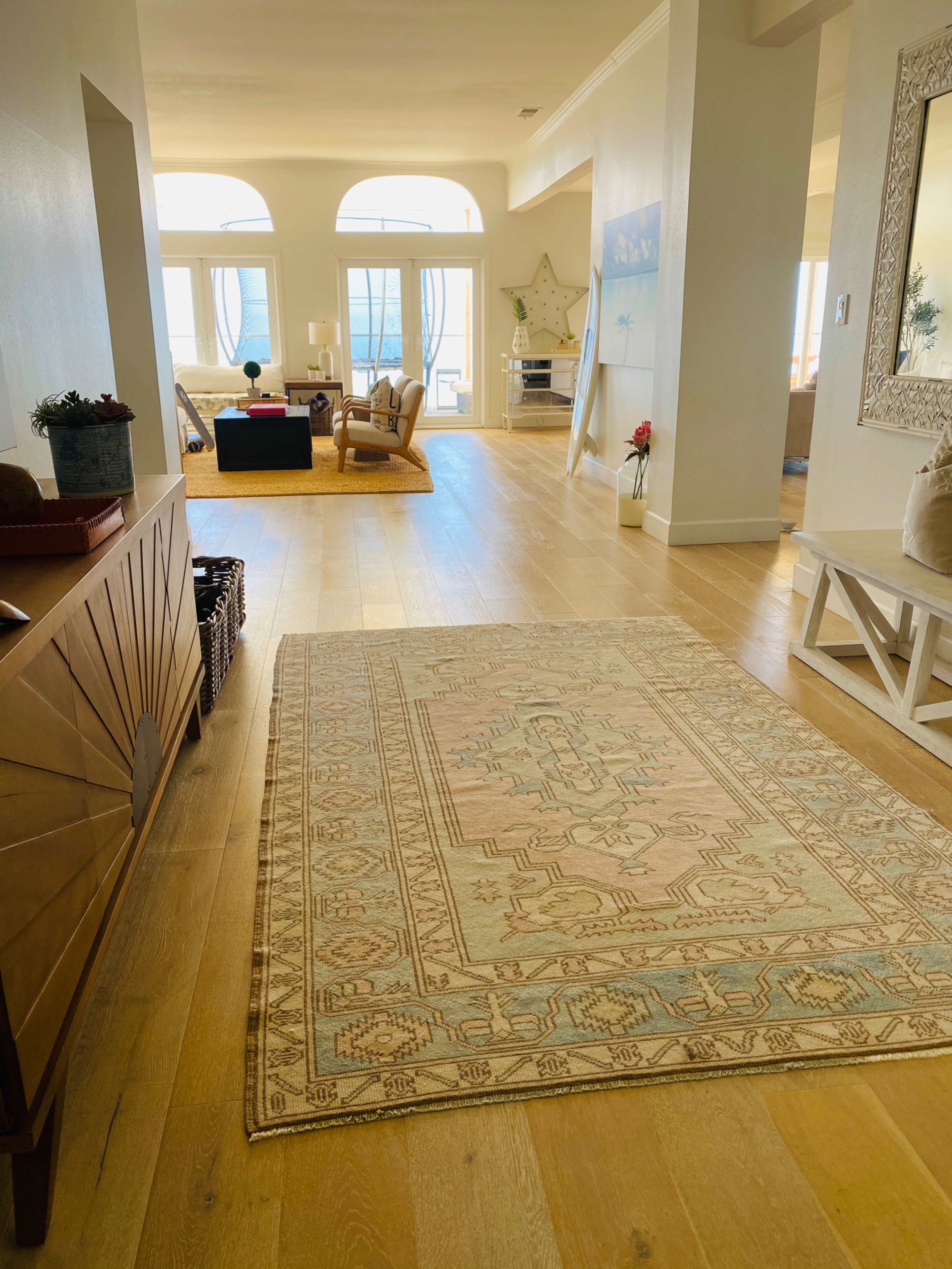 A long hallway features a decorative rug, leading to a sunlit living area with large windows and comfortable seating.