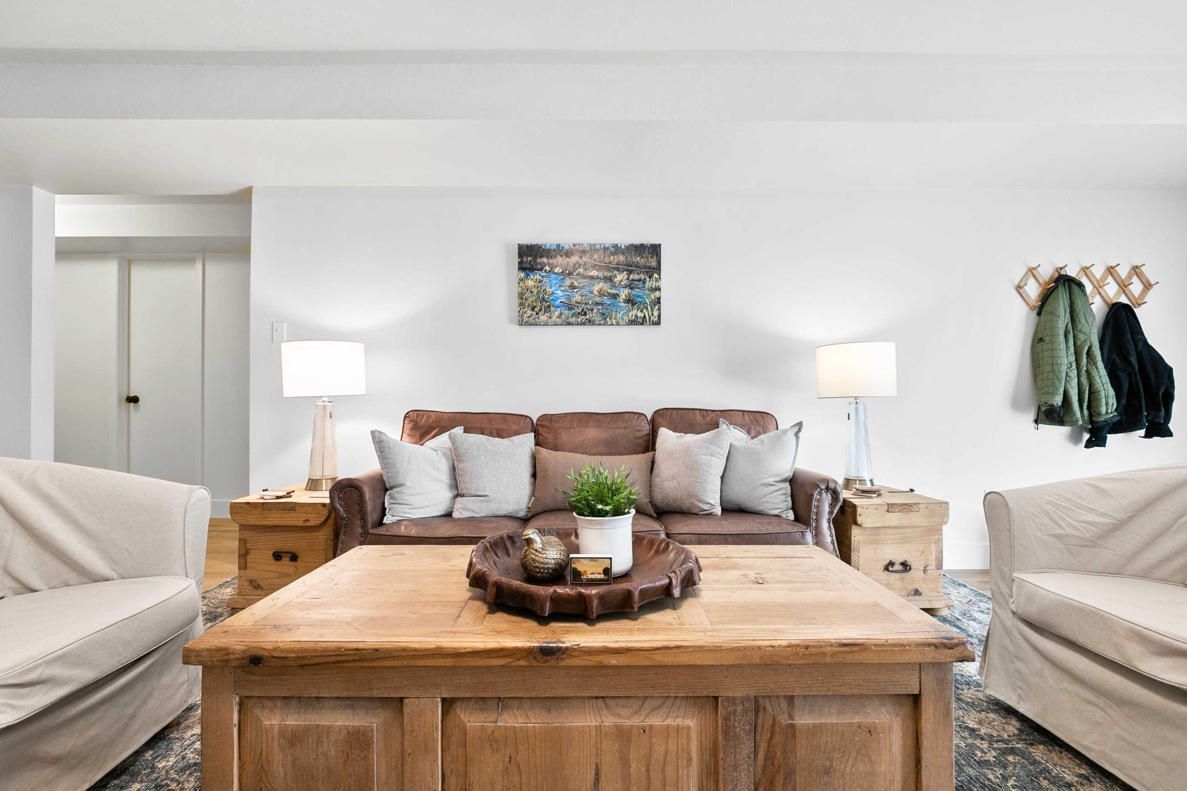 A living room features a brown leather sofa with decorative pillows, flanked by two light-colored chairs and a wooden coffee table, all set against a neutral wall and complemented by lamps and a piece of artwork.