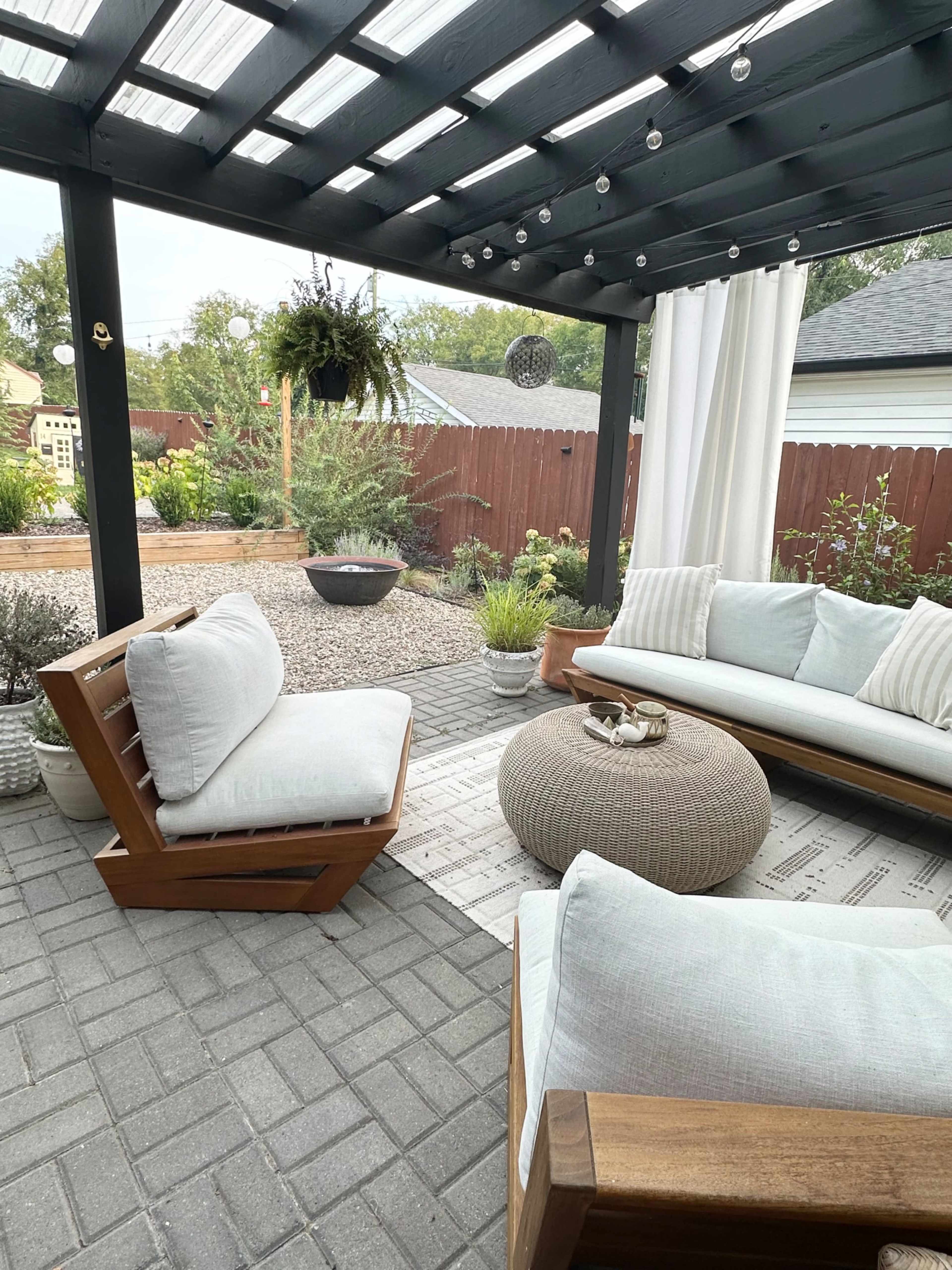 A cozy outdoor seating area with wooden furniture, a round pouf, and decorative plants under a shaded pergola.