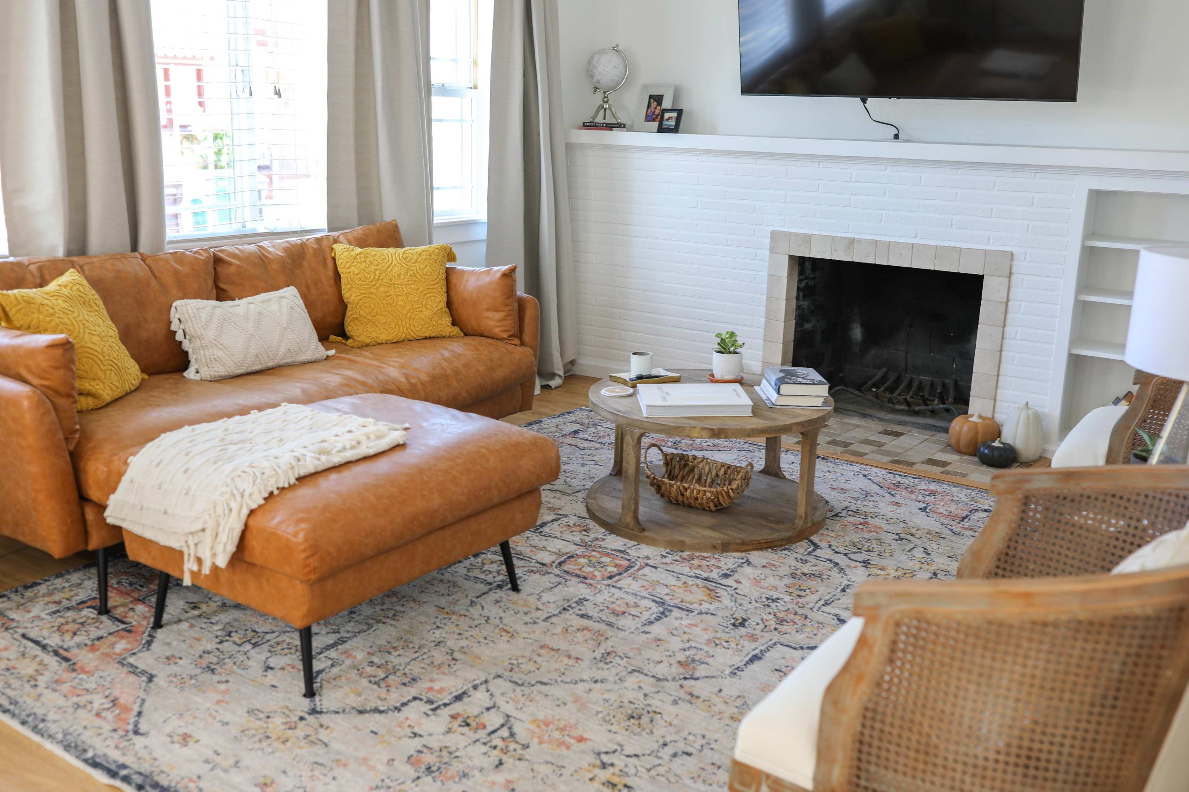 A cozy living room features a brown leather sofa, a round wooden coffee table, and a white brick fireplace with a television mounted above it.