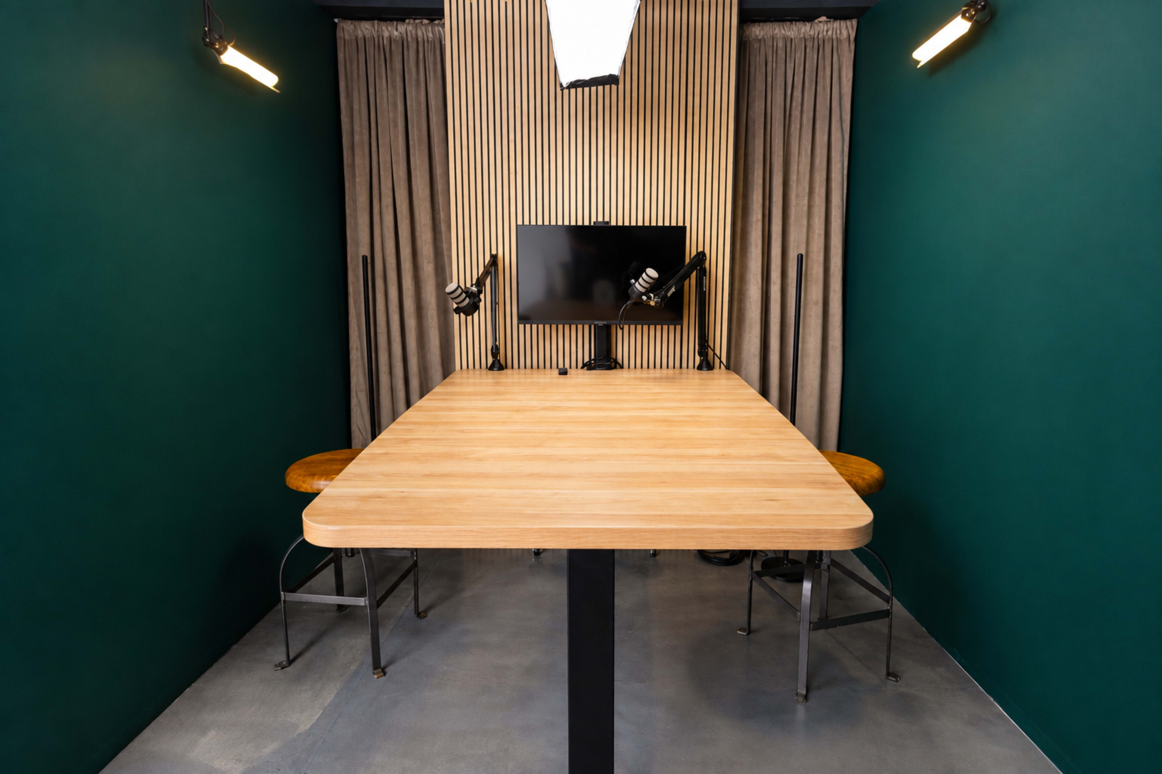 The image shows a modern, minimalistic workspace with a wooden table, two chairs, a monitor, and studio lights against a green wall and striped backdrop.
