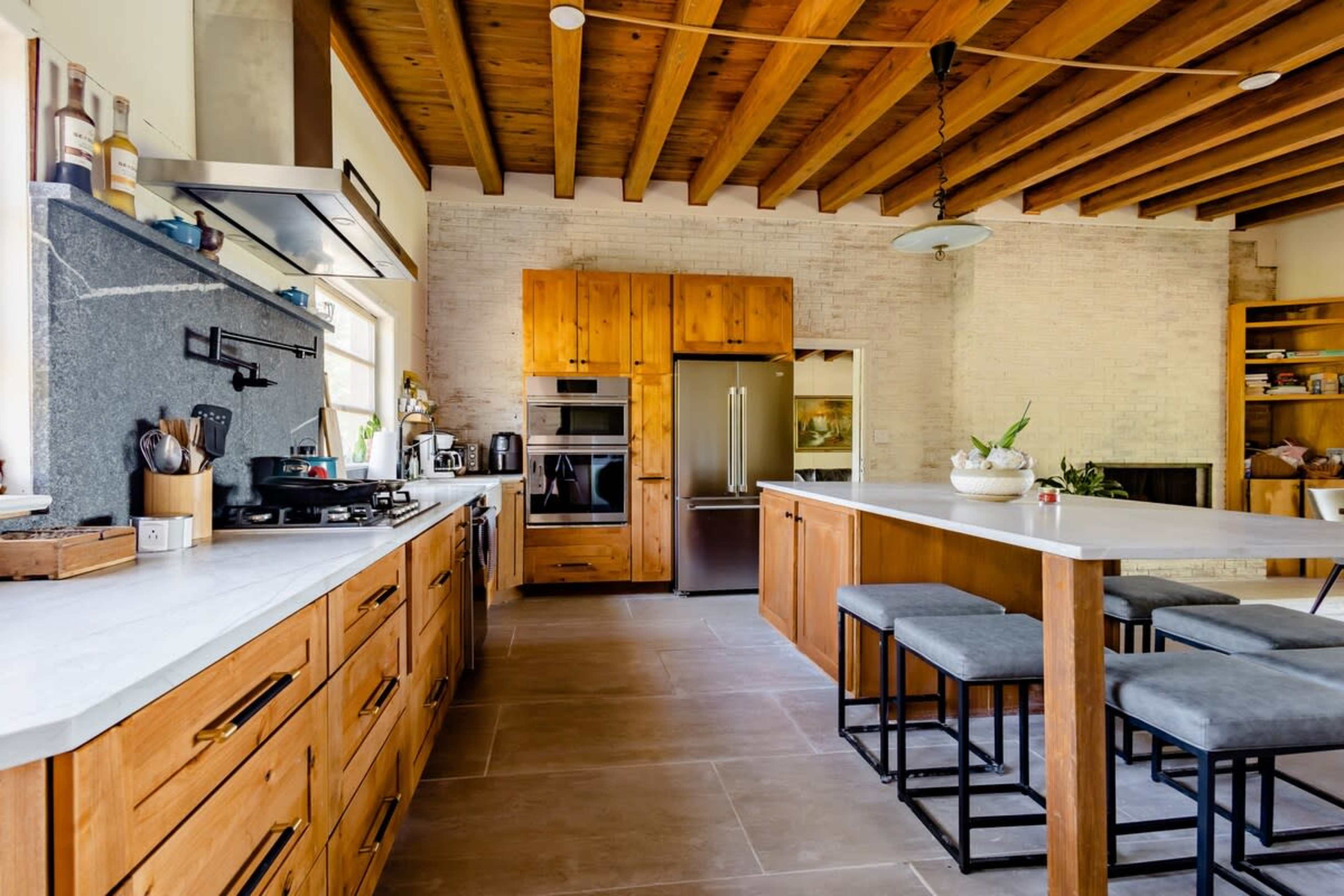 A modern kitchen features wooden cabinetry, a large island with bar stools, stainless steel appliances, and exposed wooden beams on the ceiling.
