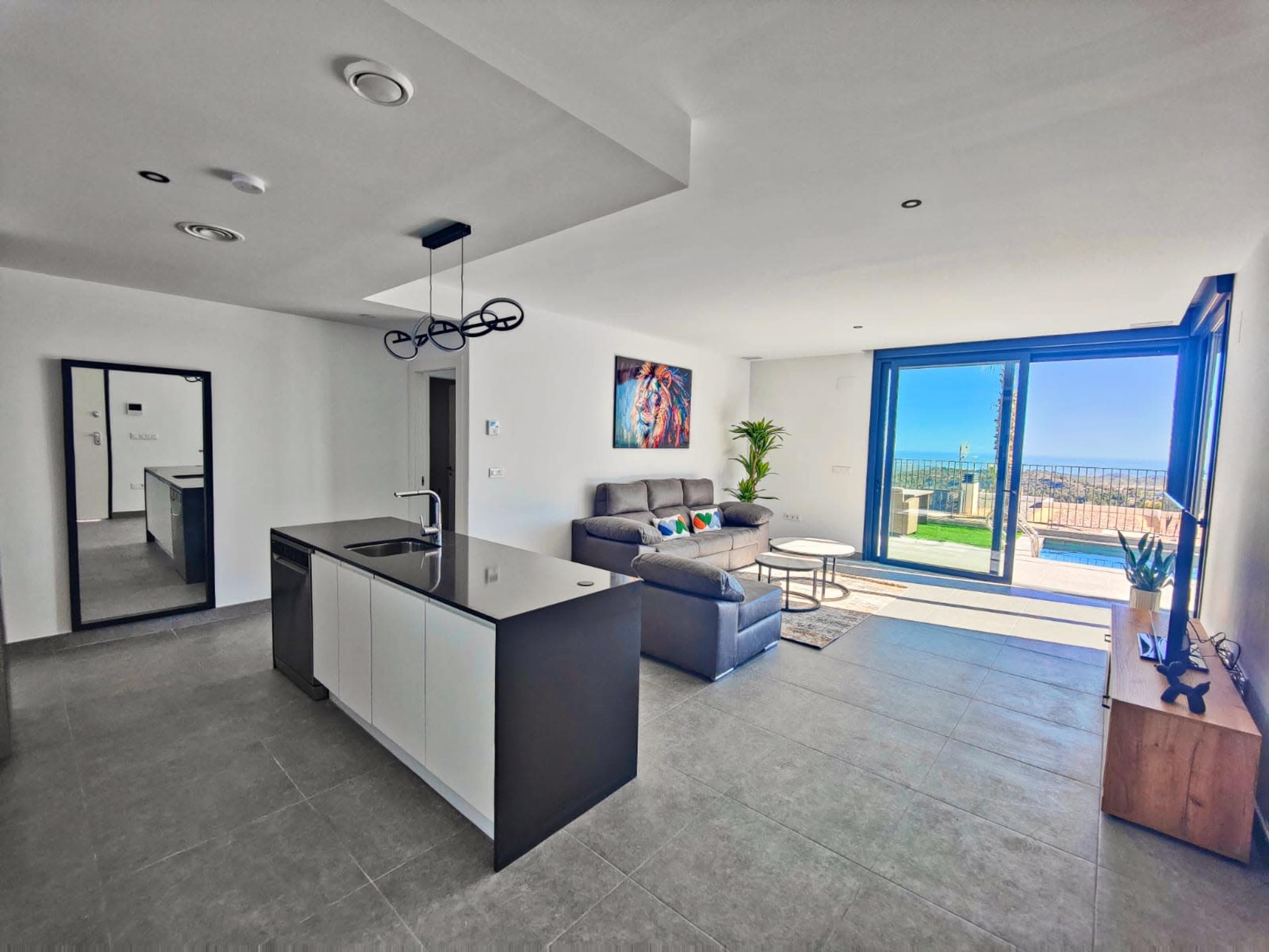 The image shows a modern living space with a kitchen island, a sofa area, a large glass door leading to an outdoor space, and a mirror on the wall.