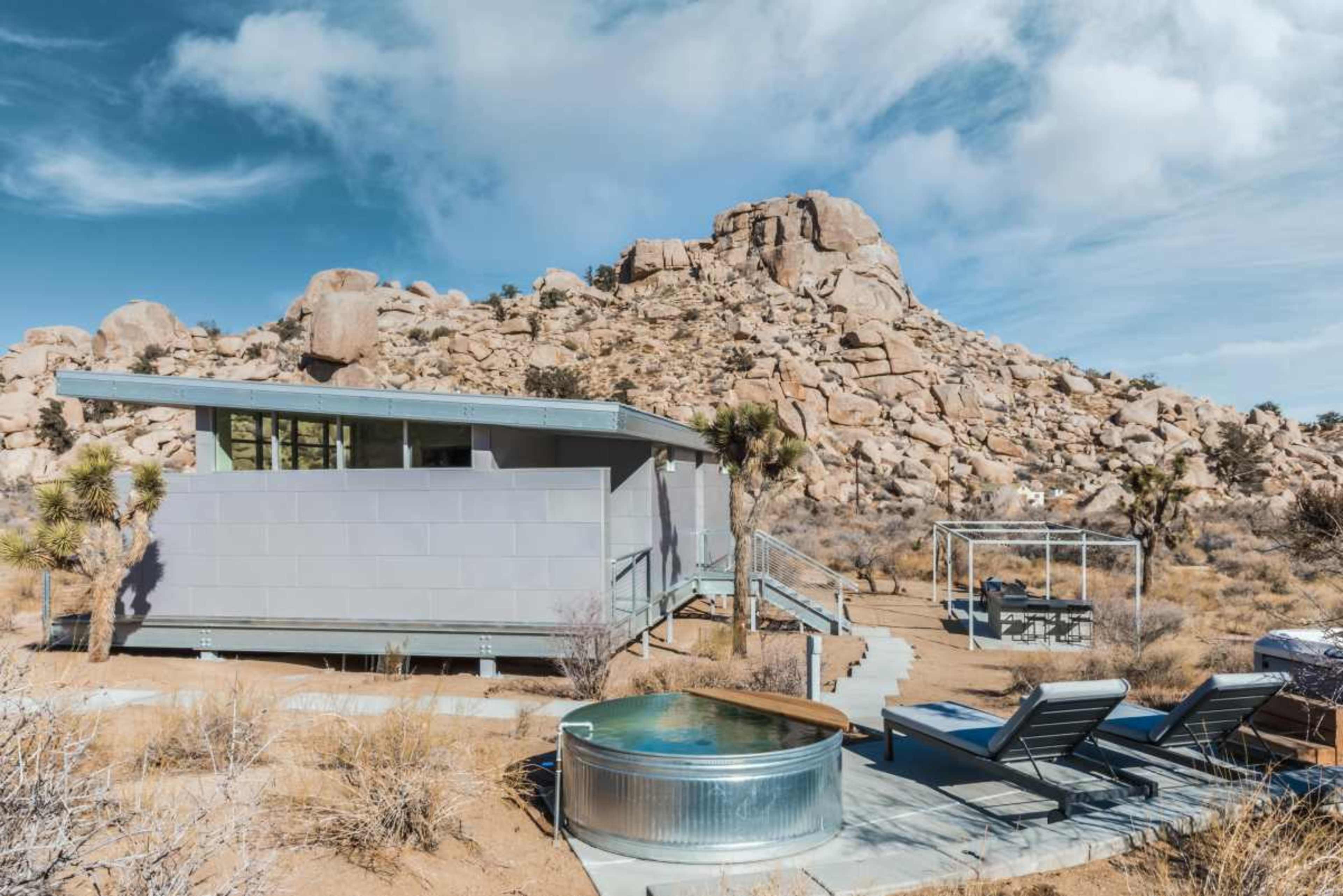 A modern, gray house with a hot tub and lounge chairs is set against a backdrop of rocky hills and desert vegetation.