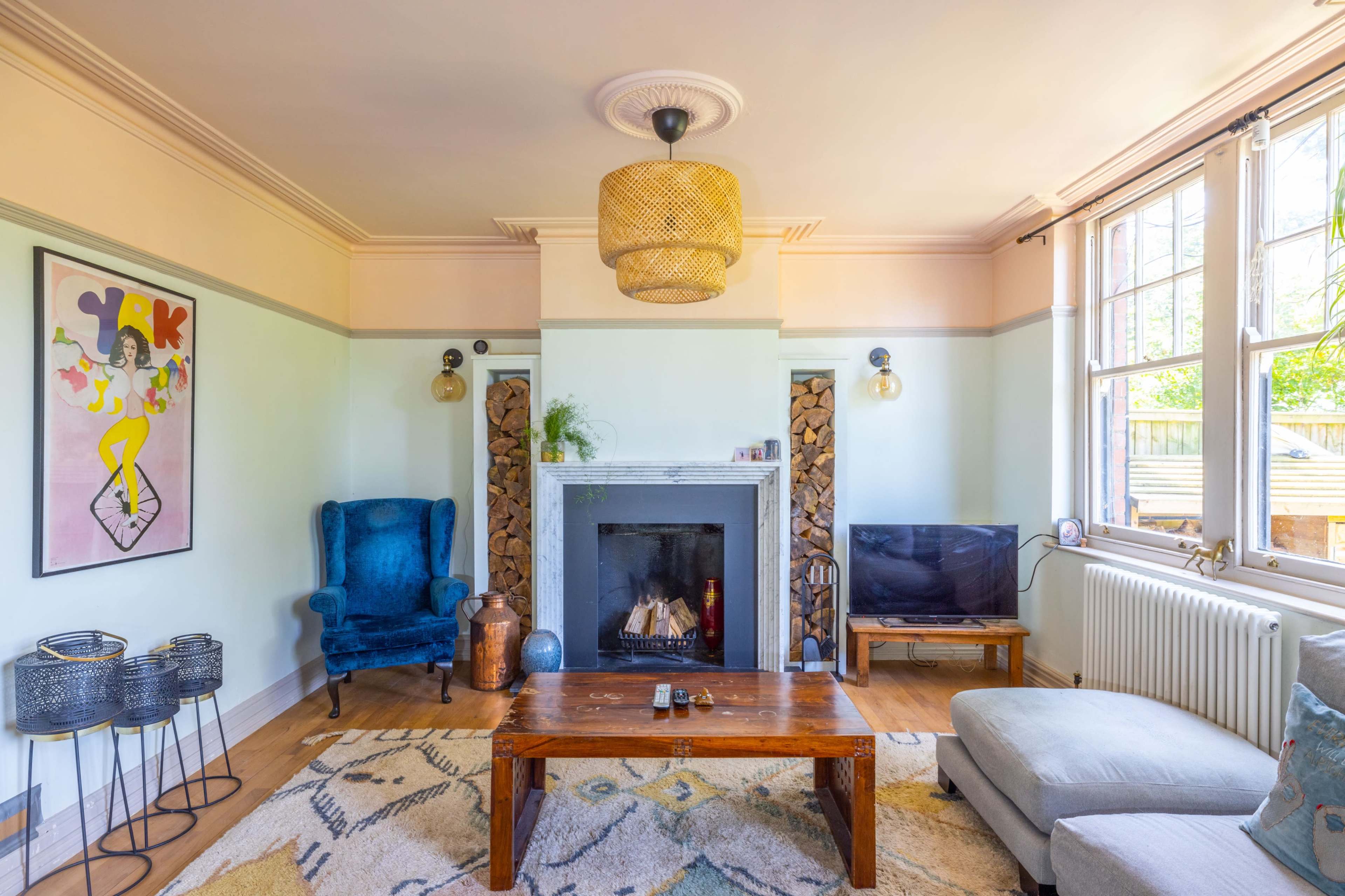A living room features a blue armchair, a wooden coffee table, a television, a fireplace with stacked wood, and large windows allowing natural light.