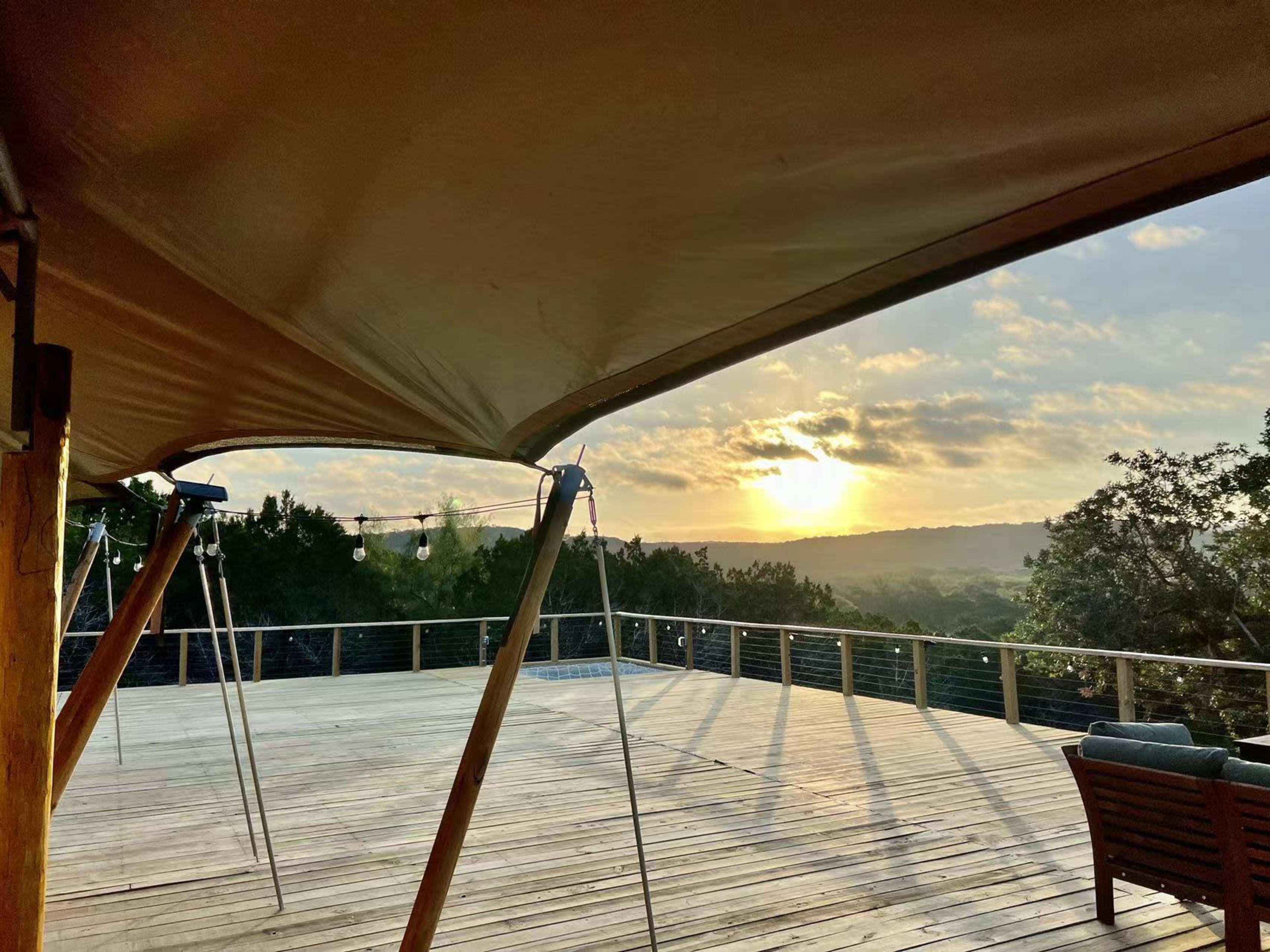 A wooden deck with a canopy and outdoor seating overlooks a valley at sunset.