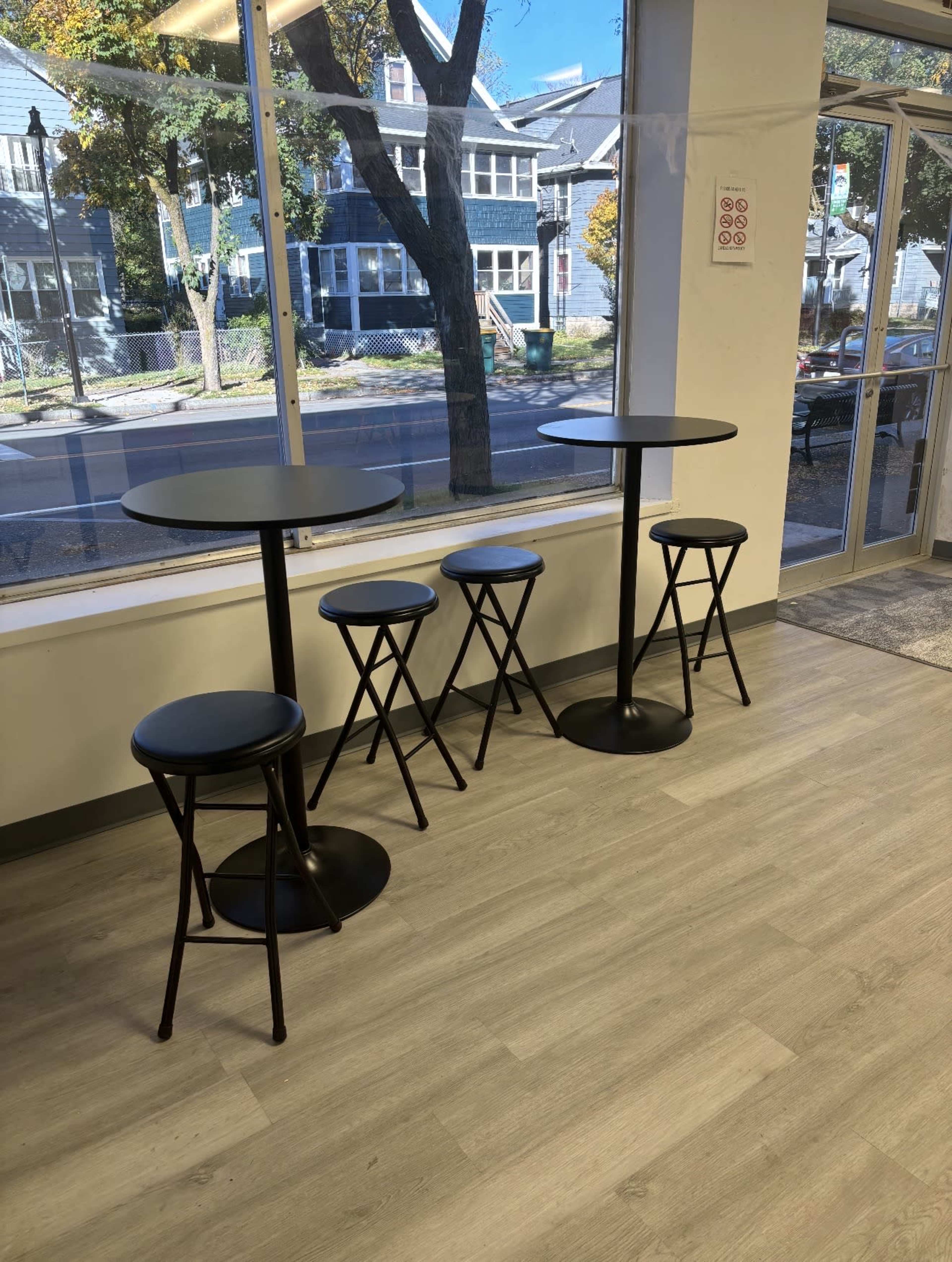 The image shows four black high-top tables with matching stools arranged near a large window, overlooking a street lined with trees and houses.