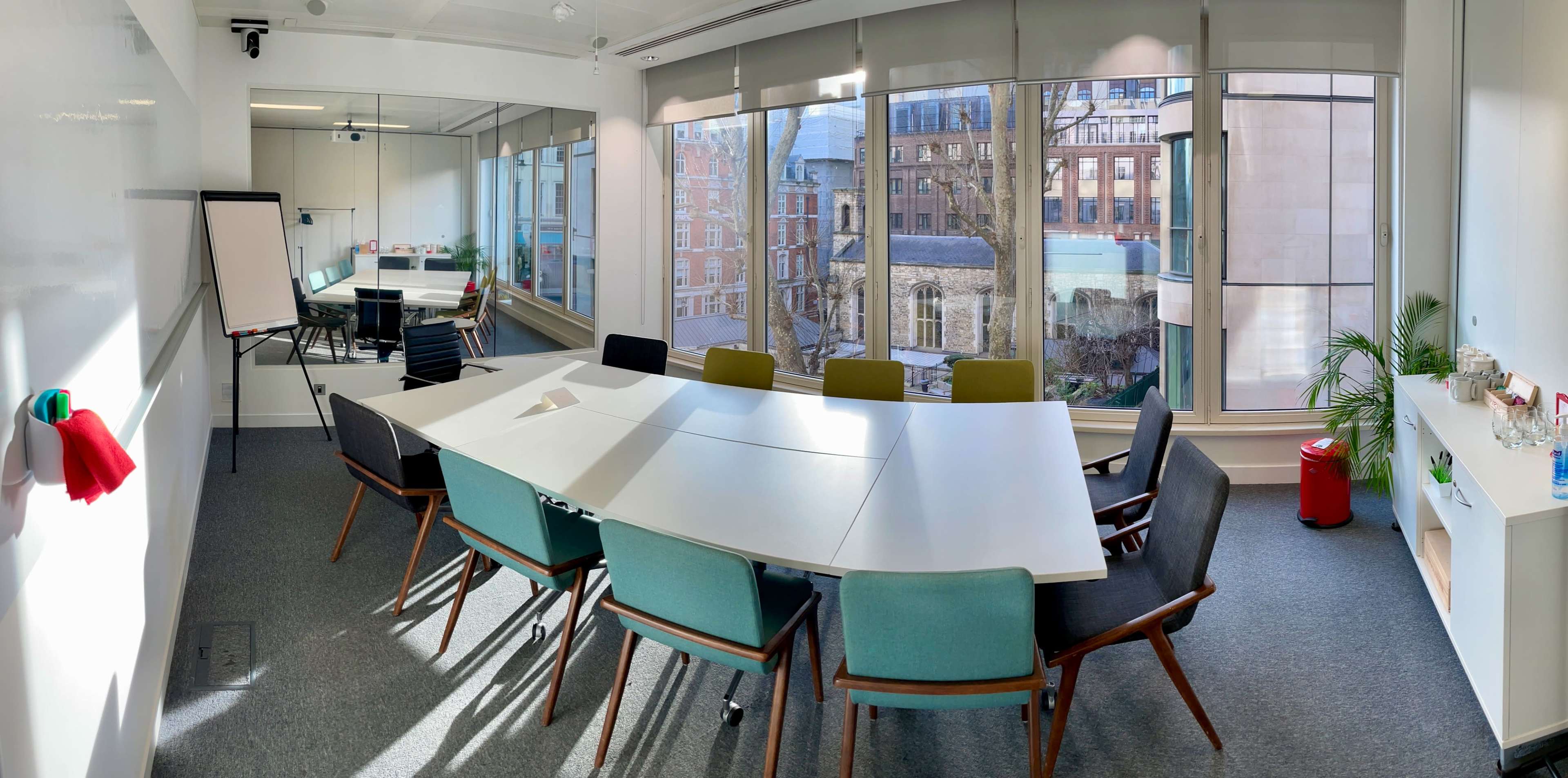 A modern conference room features a large white table surrounded by colorful chairs, with large windows overlooking an urban courtyard.