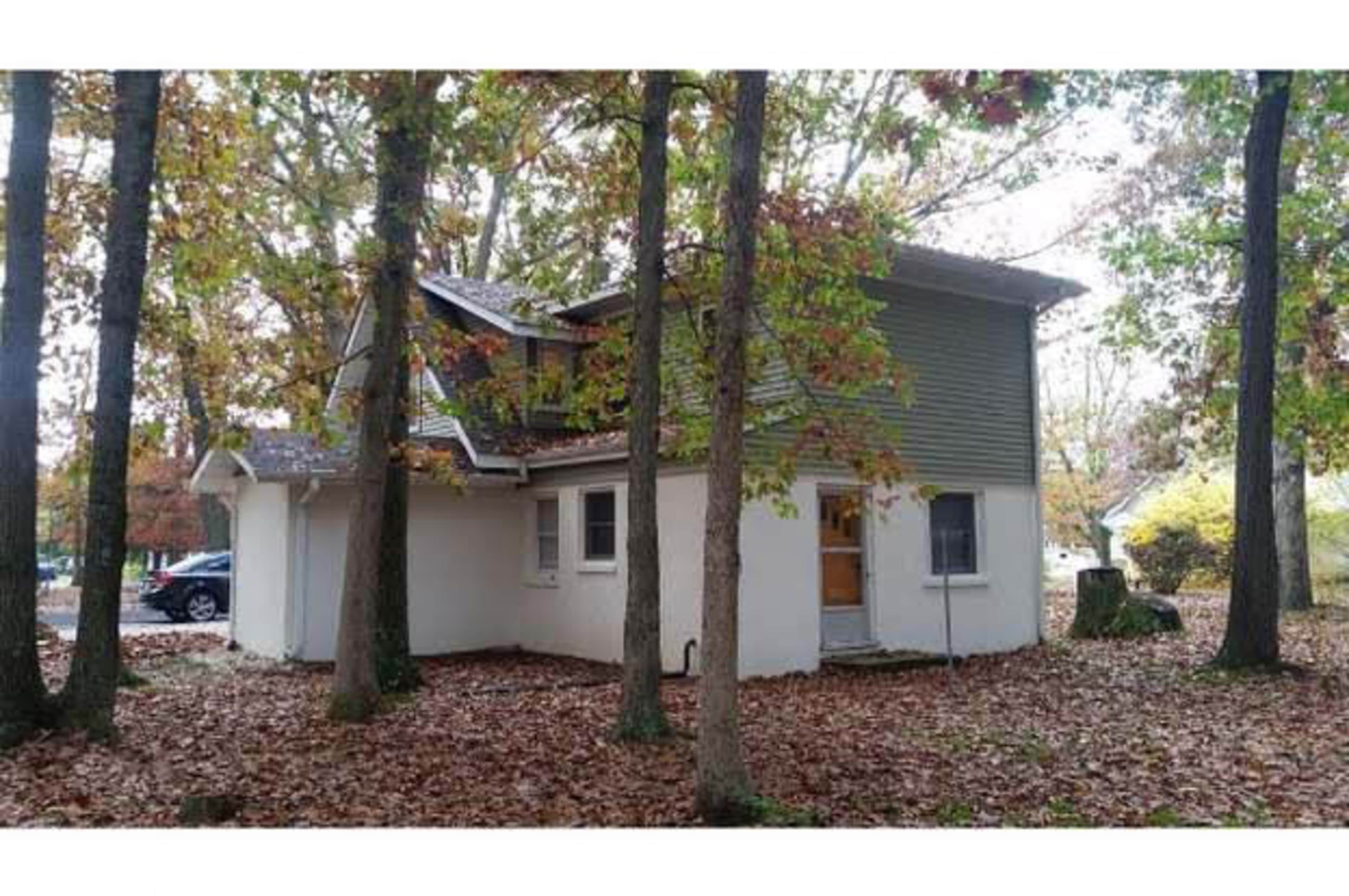 A two-story house with a green and gray exterior is surrounded by trees and fallen leaves in a wooded area.