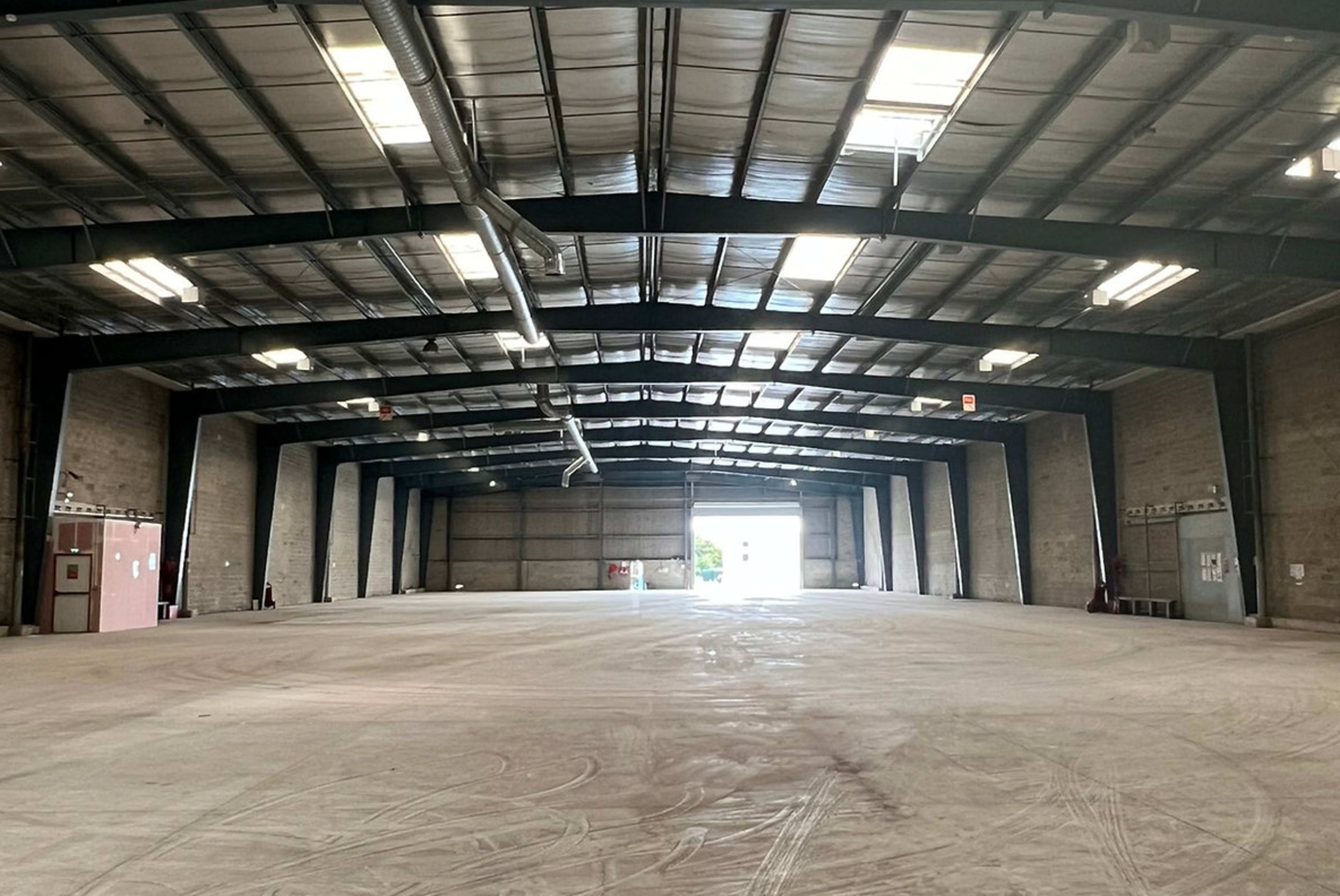 An empty industrial warehouse features a high ceiling with exposed beams and large overhead lighting, illuminated by natural light from the entrance.