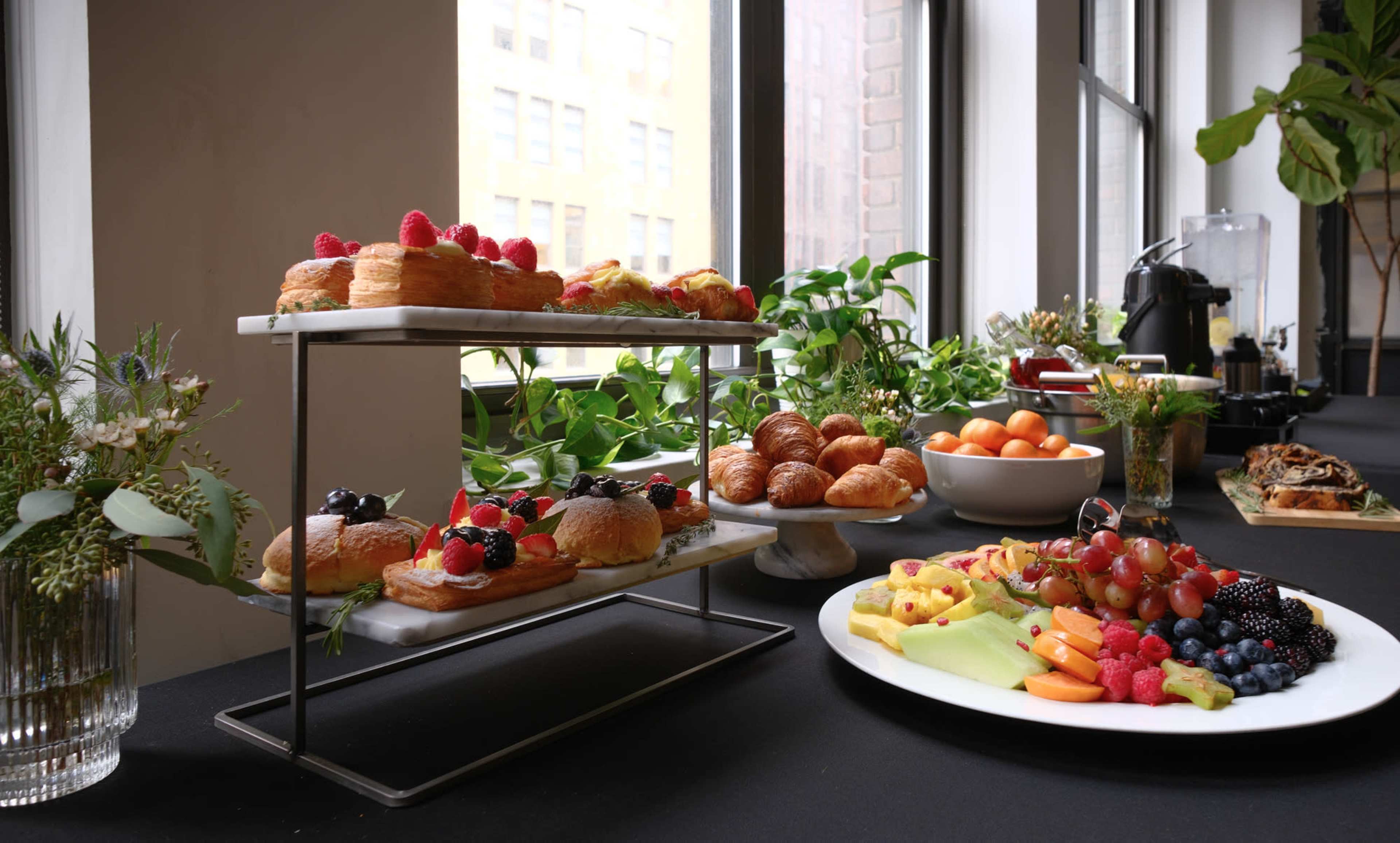 A variety of pastries and fresh fruit are displayed on a multi-tiered stand and a white plate on a table near large windows.