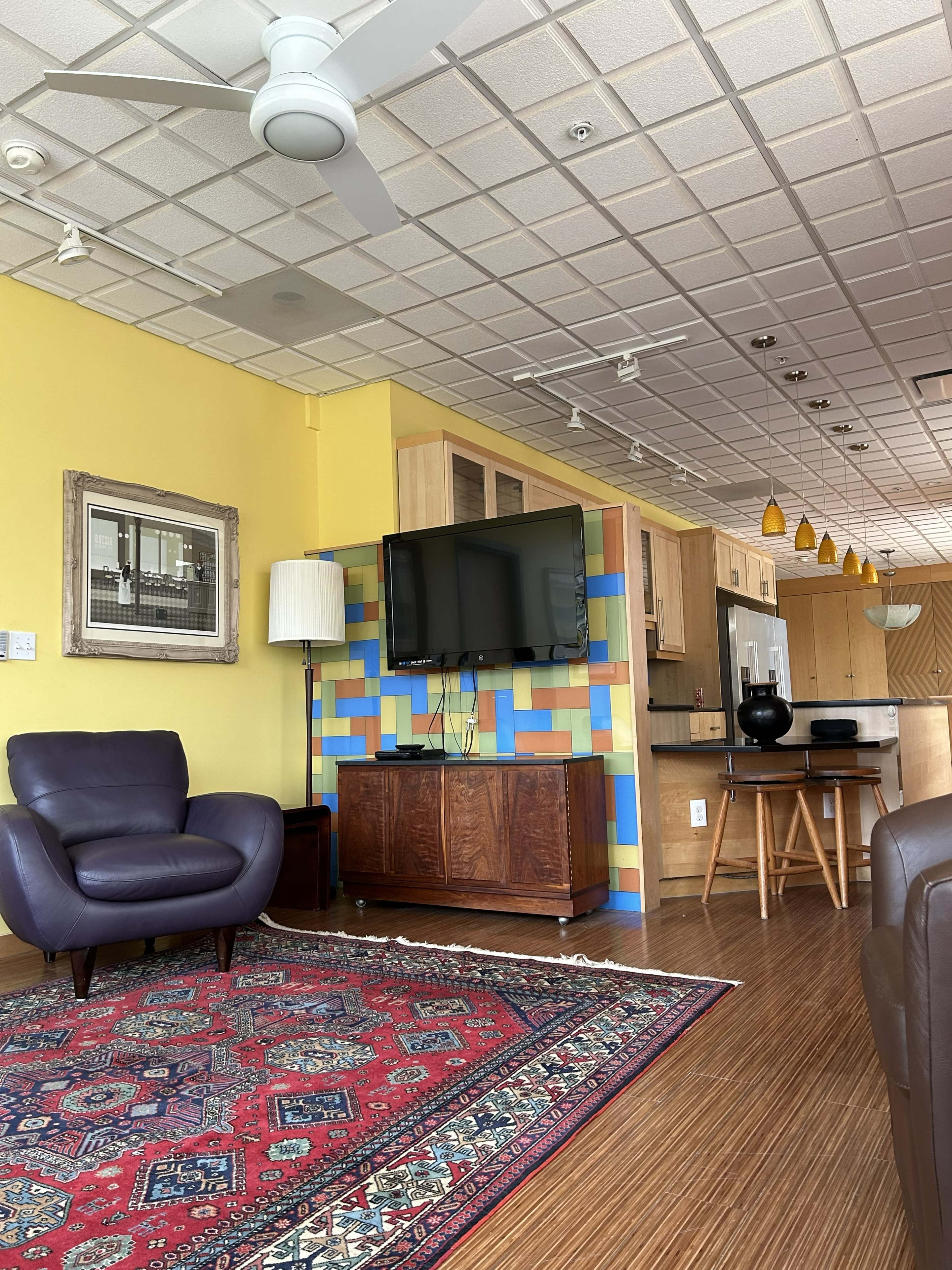 The image shows a cozy living area with a purple armchair, a wooden TV stand, a flat-screen television on the wall, and a patterned rug on a wooden floor.