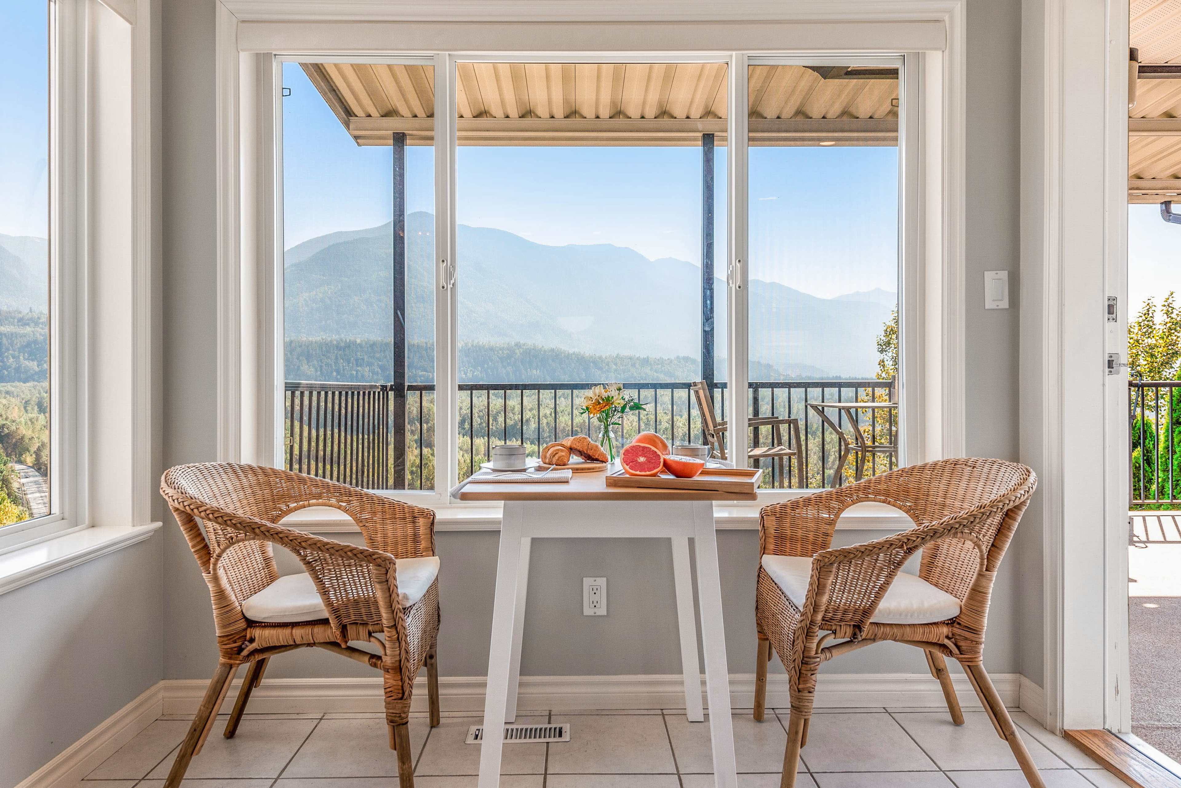 A small dining area features a table with a fruit platter and two wicker chairs, overlooking a mountain view through large windows.