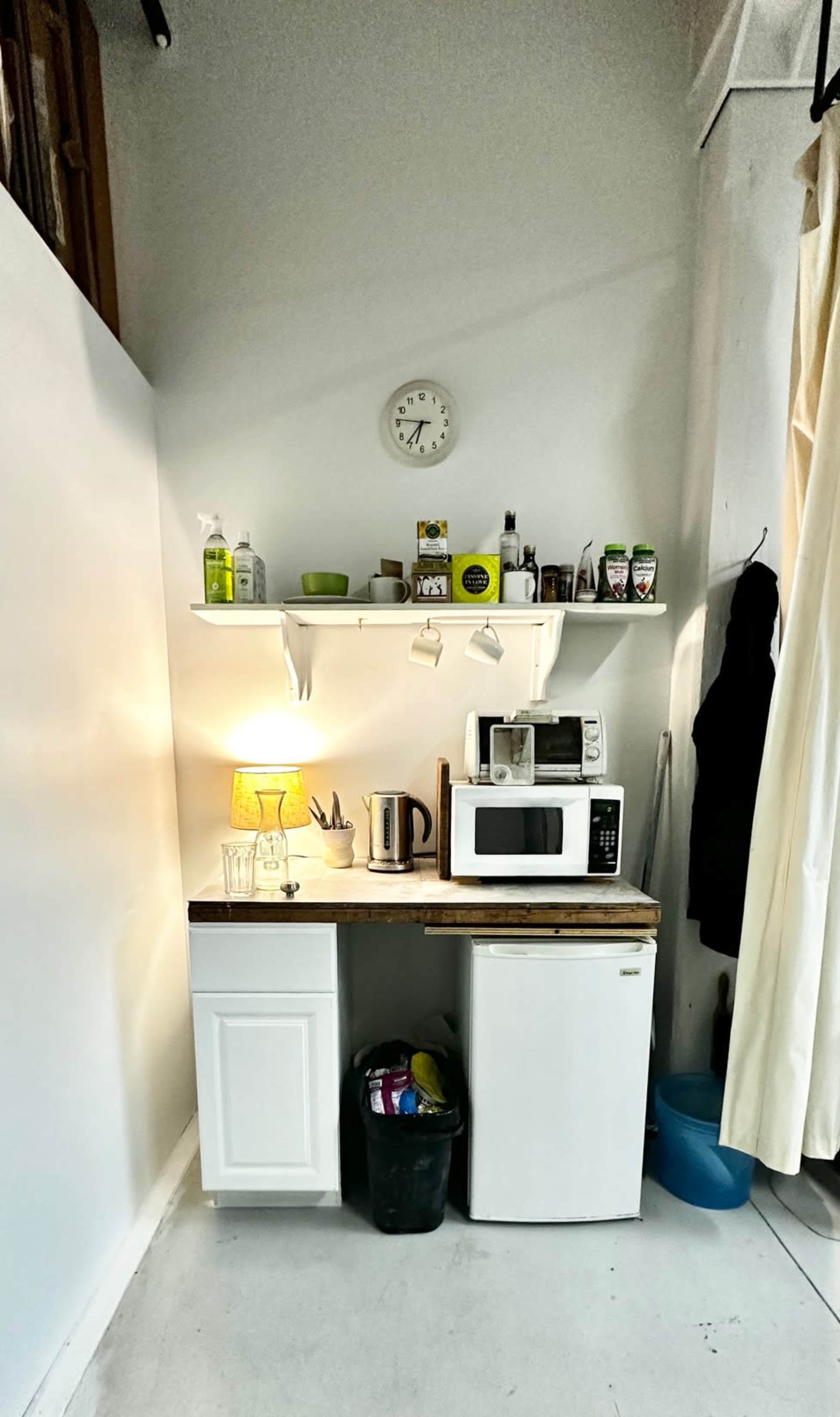 The image shows a small kitchen area with a white shelf above a wooden table, featuring a microwave, kettle, light, and various kitchen supplies, alongside a small fridge and a trash can.