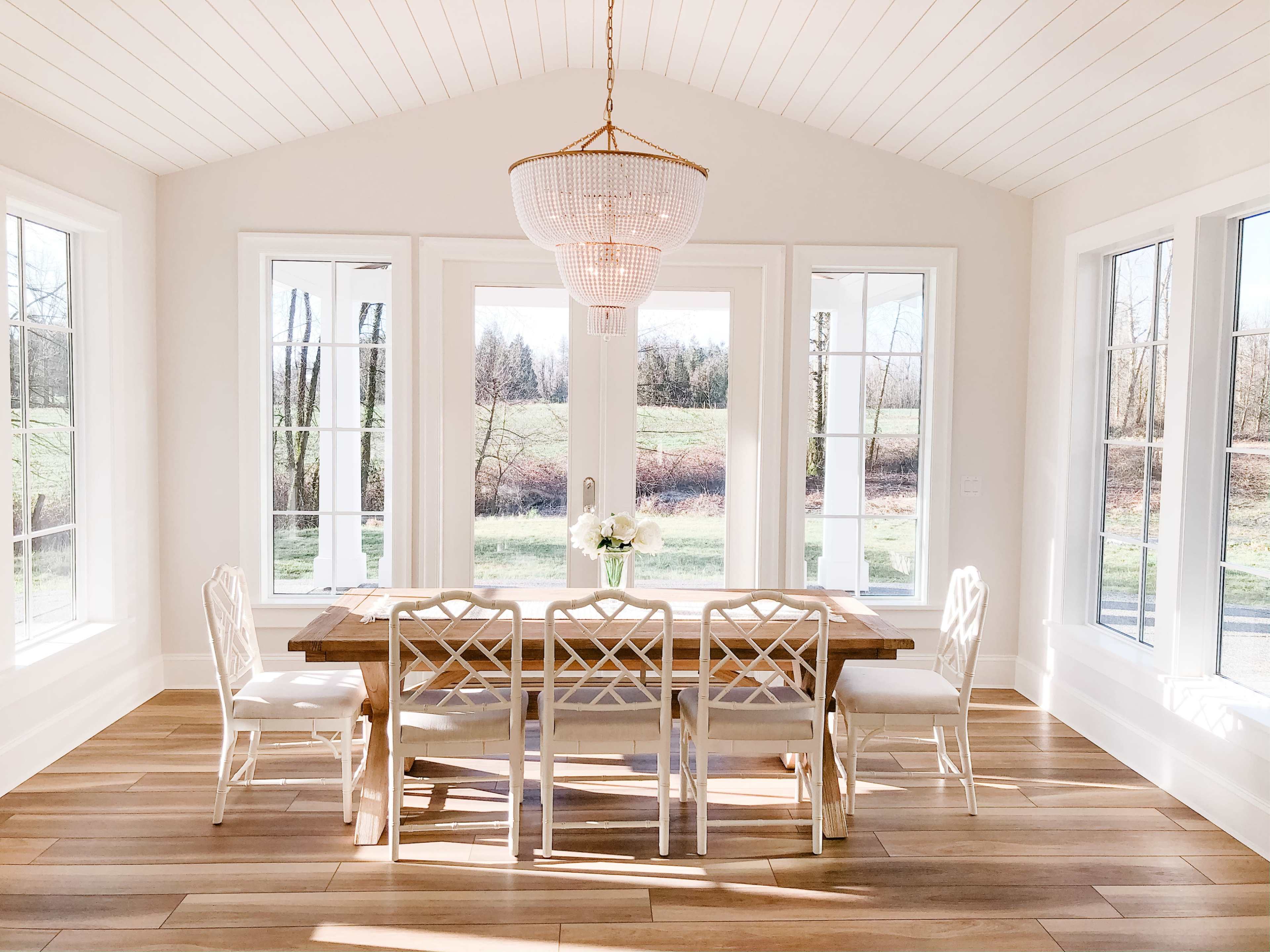 A bright dining area features a wooden table surrounded by six chairs, with large windows providing a view of a green field.