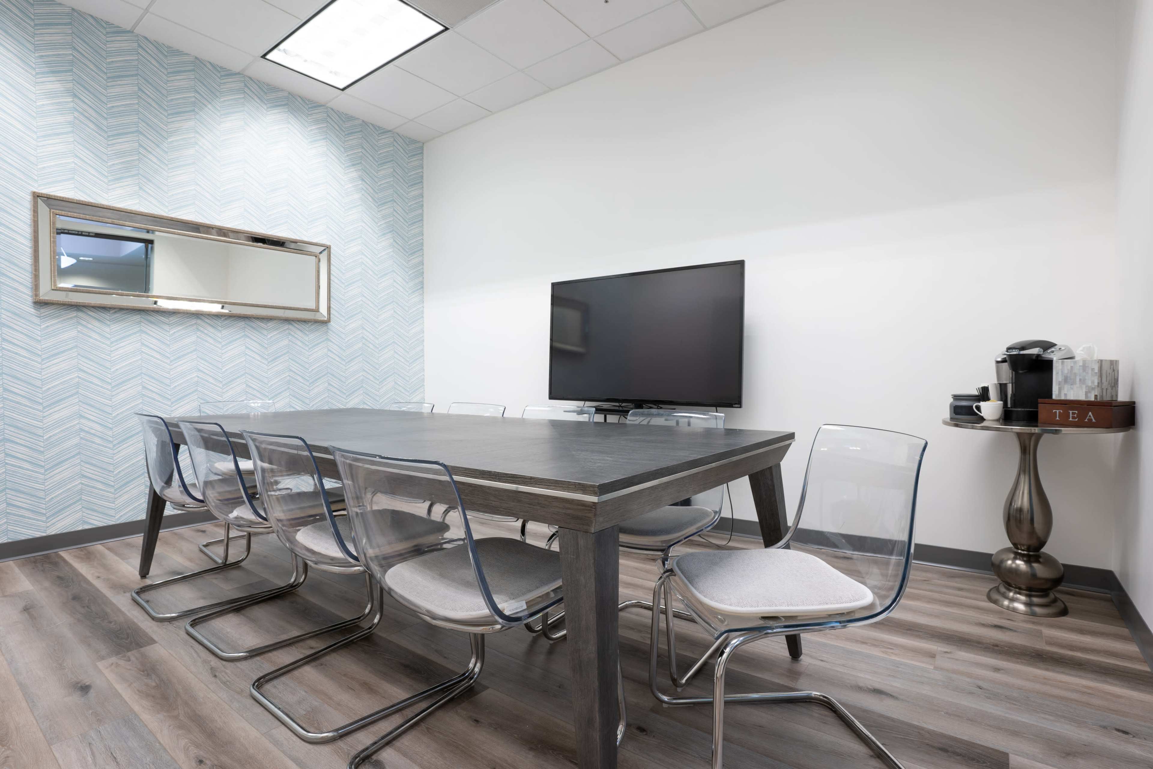 The image shows a conference room with a long wooden table surrounded by transparent chairs, a large wall-mounted television, and a coffee station on a side table.