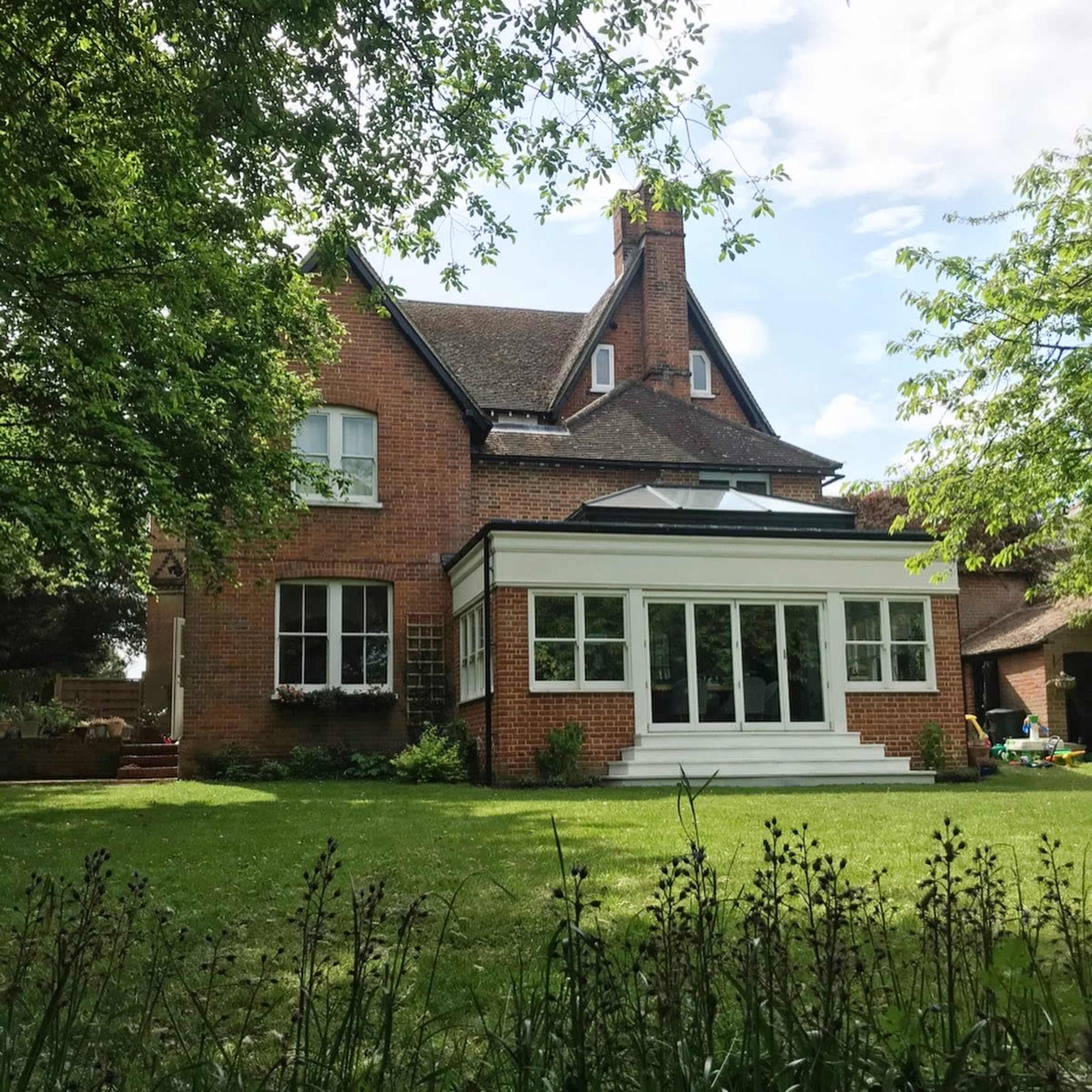 A brick house with a white conservatory and large windows sits surrounded by greenery on a sunny day.