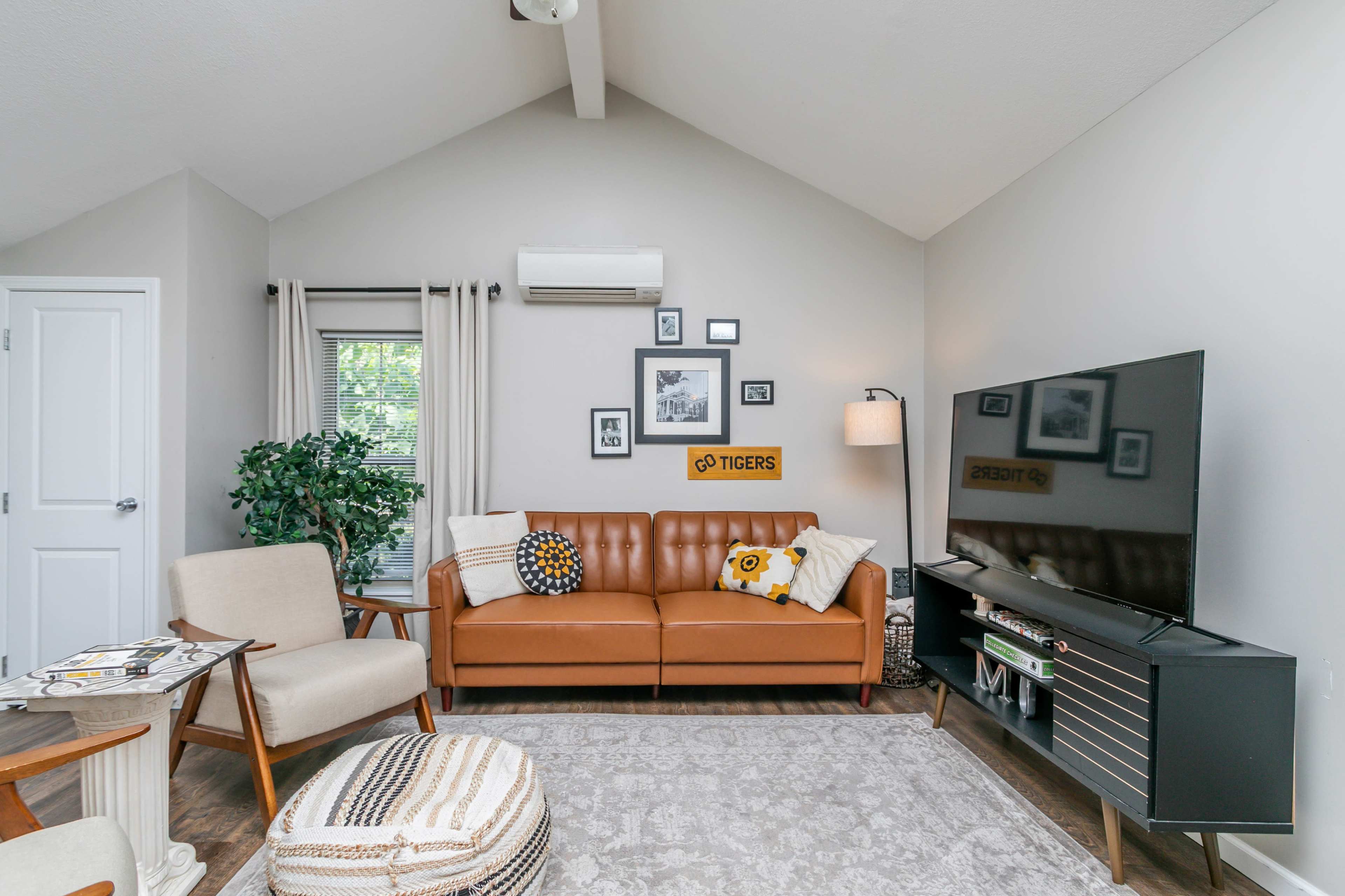 The scene depicts a cozy living room with a brown leather couch, a TV stand, and decorative framed pictures on the wall.