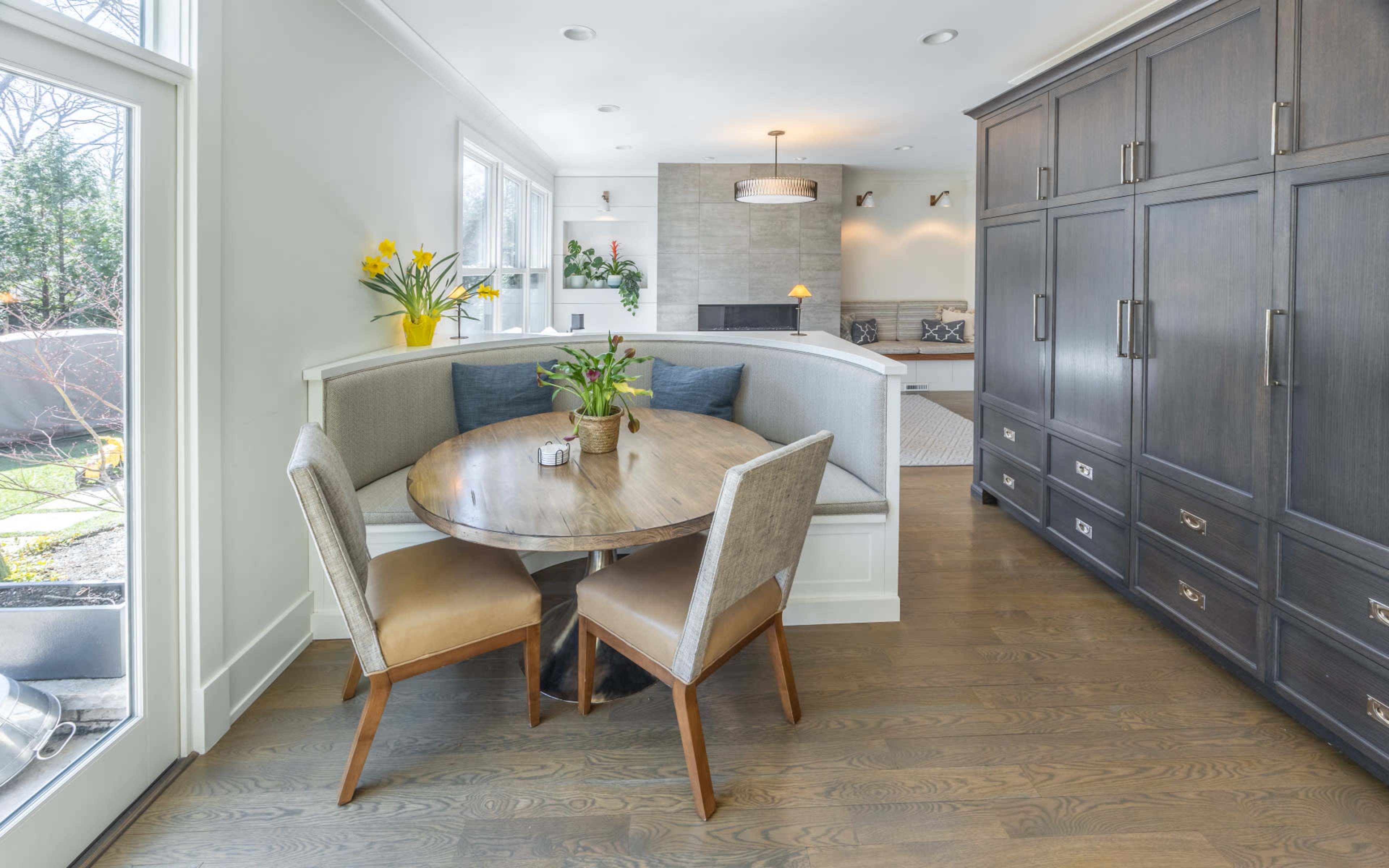 The image shows a modern dining nook with a round wooden table, two upholstered chairs, and built-in seating, adjacent to a dark cabinetry wall and a living room with a fireplace in the background.