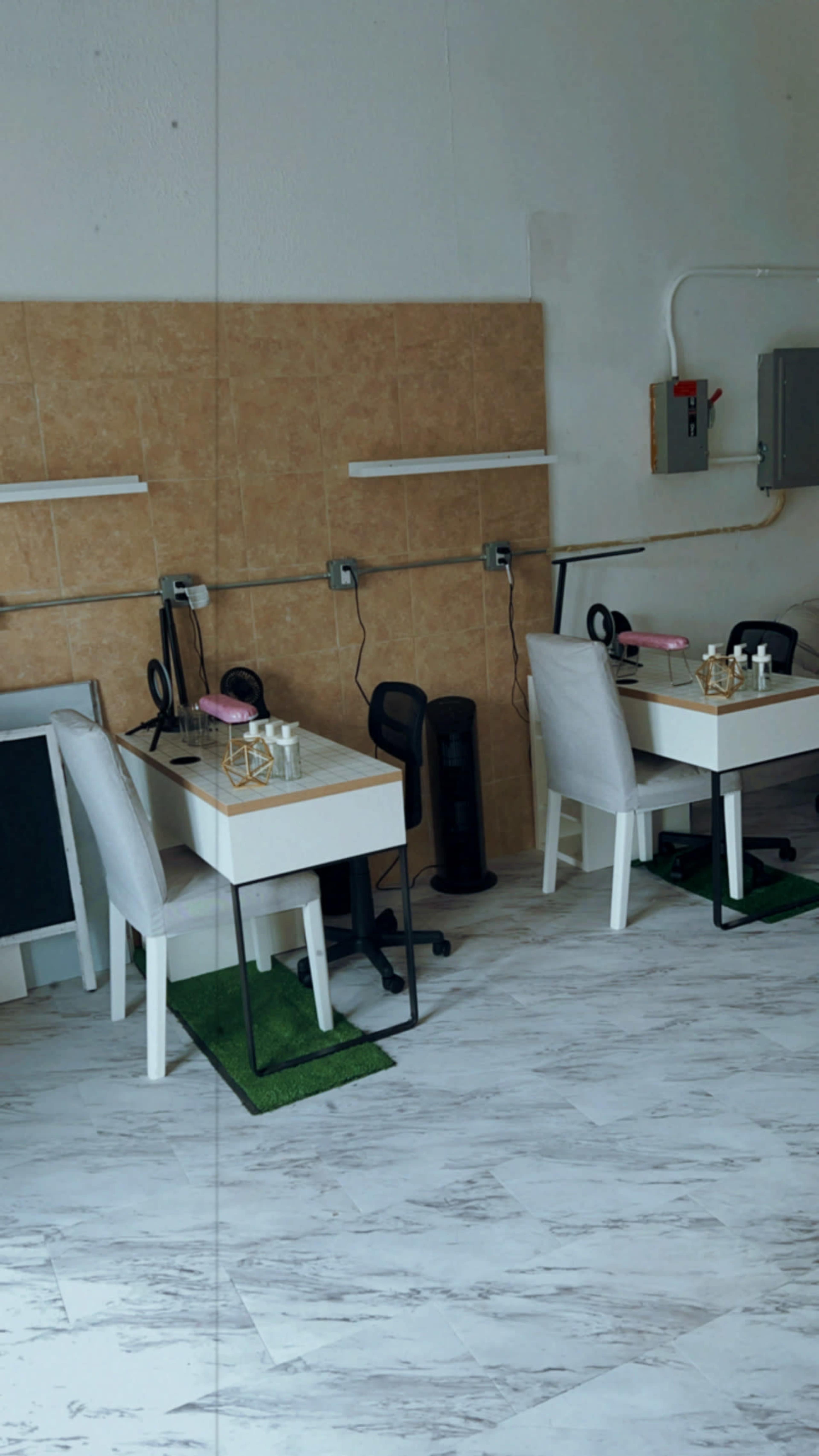 The image shows a minimalist workspace with two white desks and chairs arranged in a clean, well-lit room featuring a tiled wall and electrical panel.