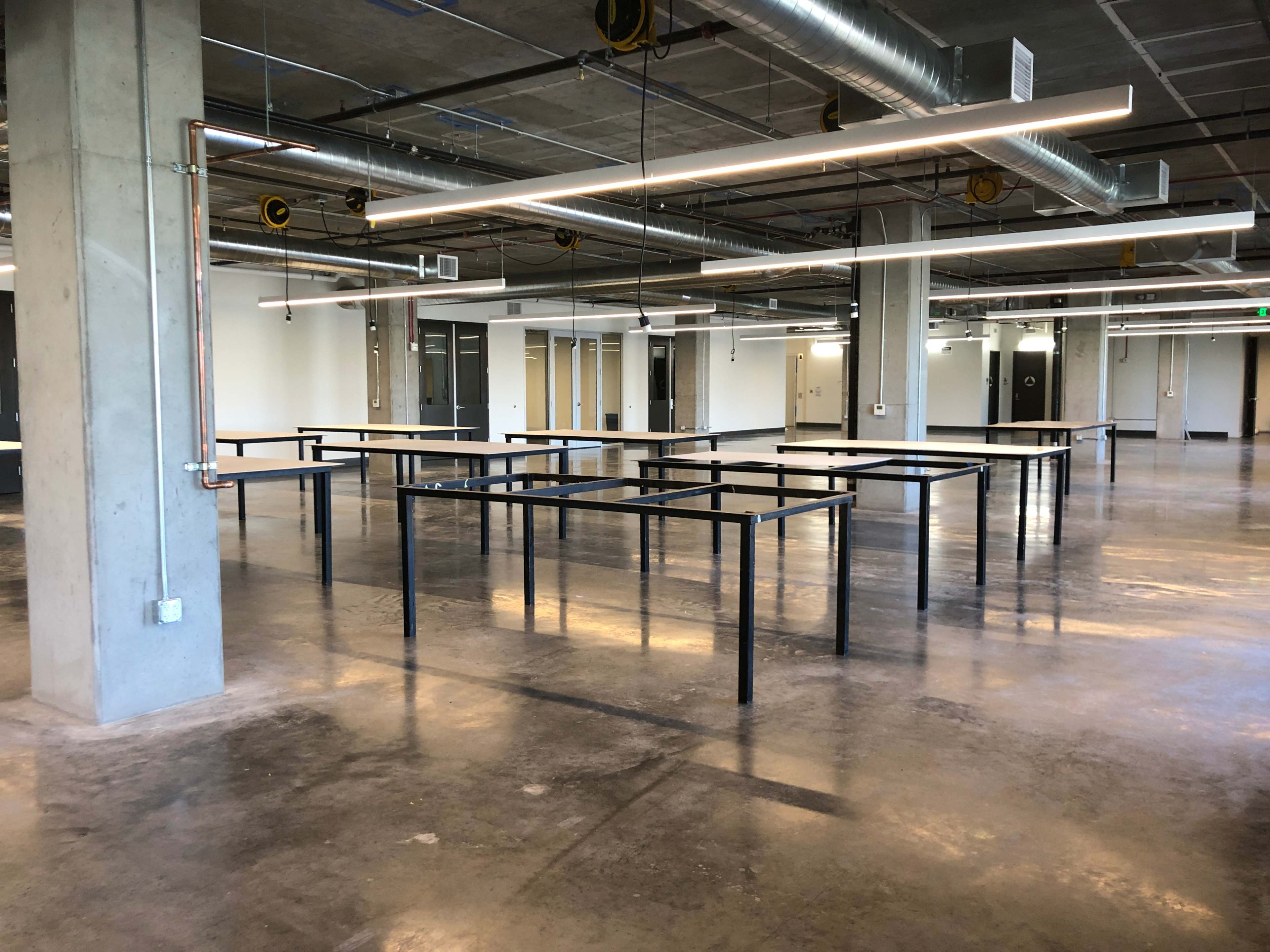 An empty room with concrete floors and several black metal tables arranged in a grid pattern under overhead lights.
