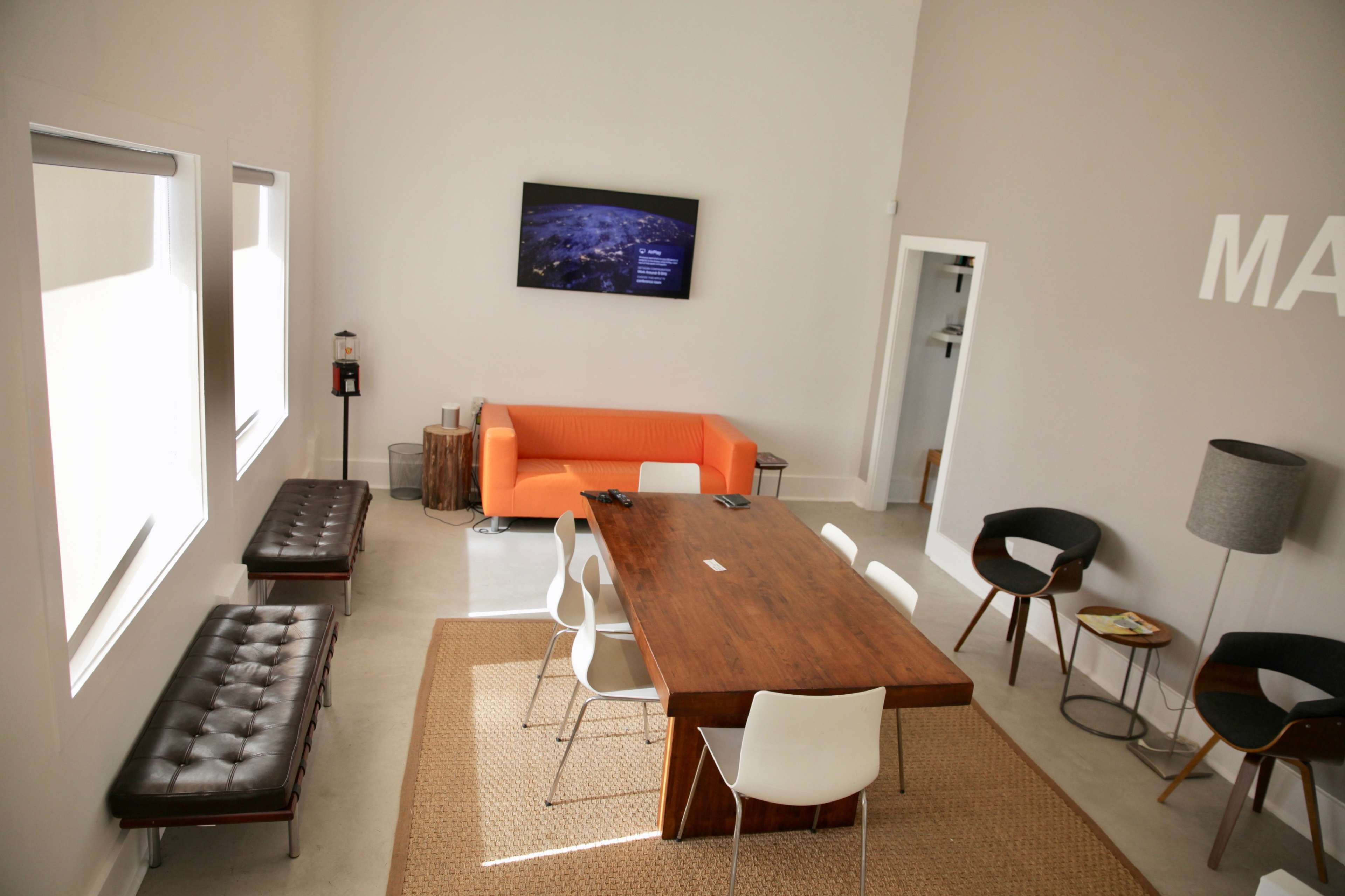 A modern meeting room features a large wooden table surrounded by white chairs, an orange sofa, and a TV mounted on the wall.