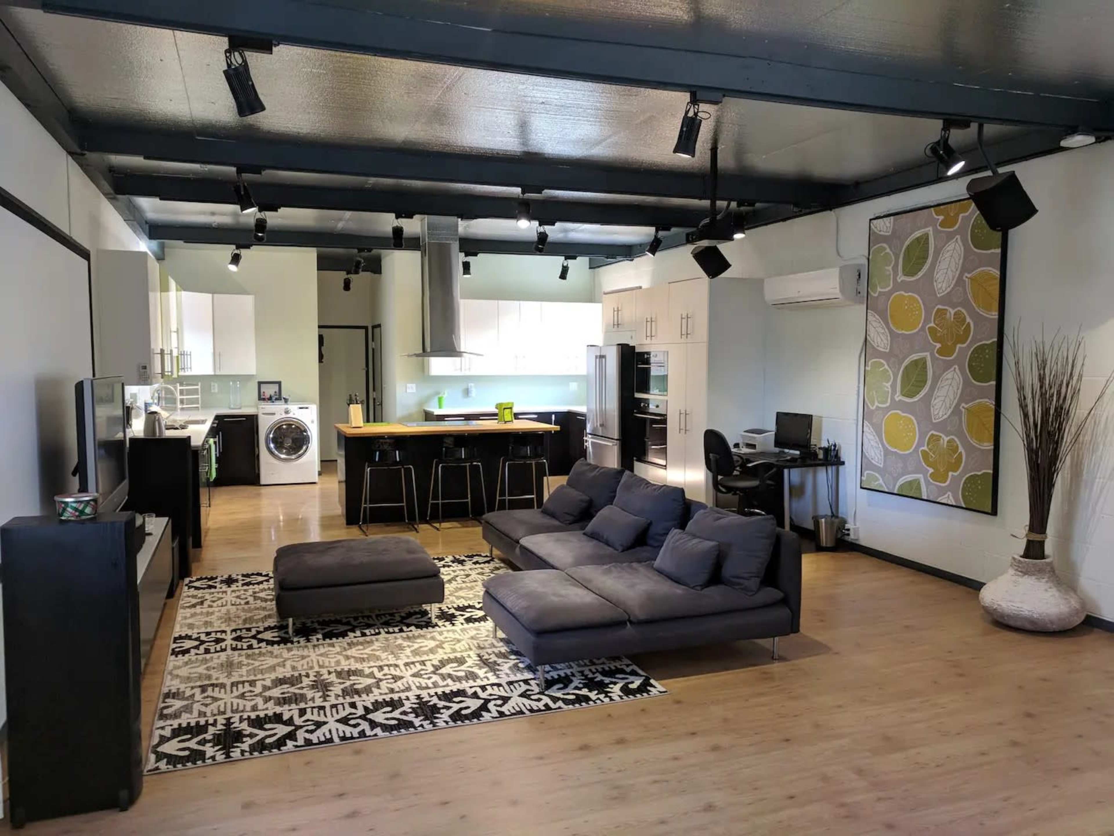 A modern living area features a black sectional sofa and a rug, with a kitchen and laundry area visible in the background.