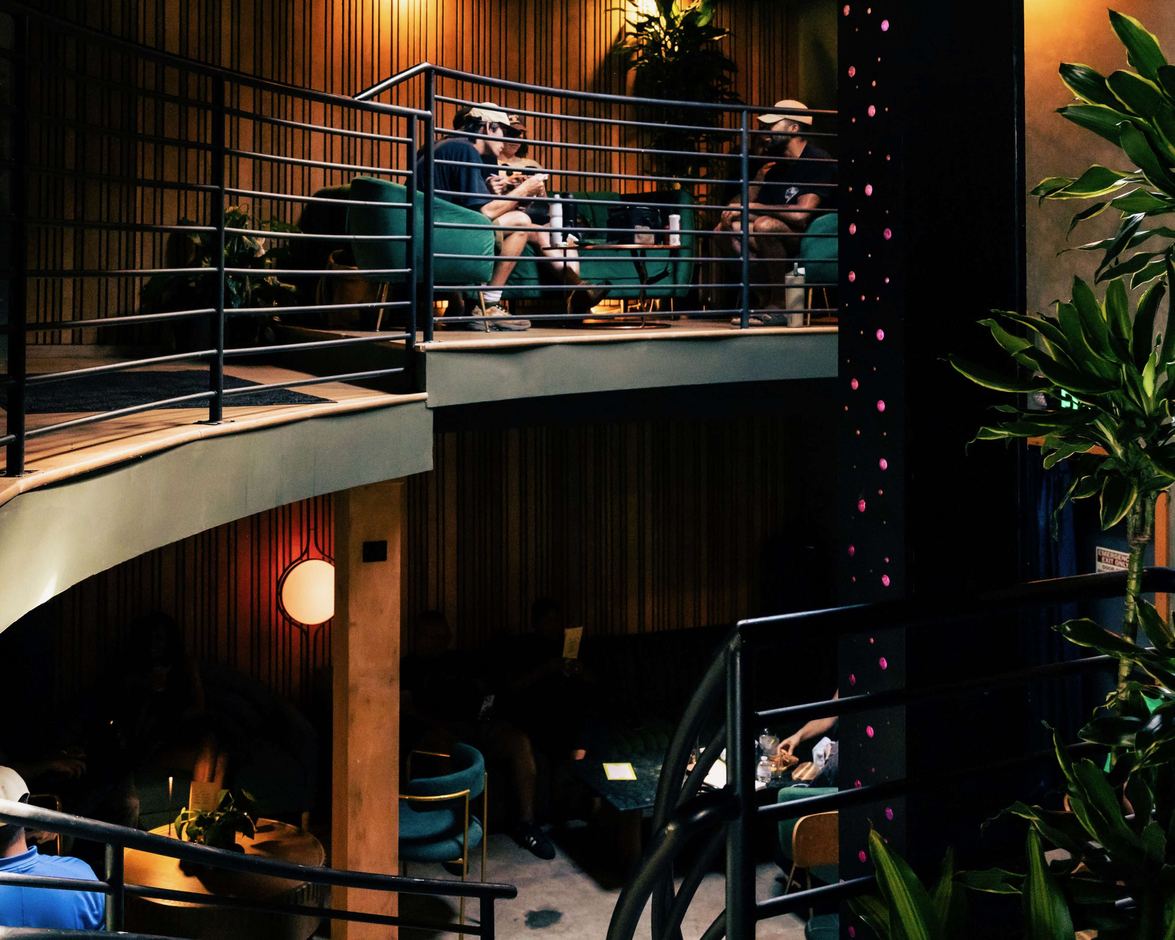 The scene depicts a modern, multi-level lounge with patrons seated on green chairs and plants positioned around the space.
