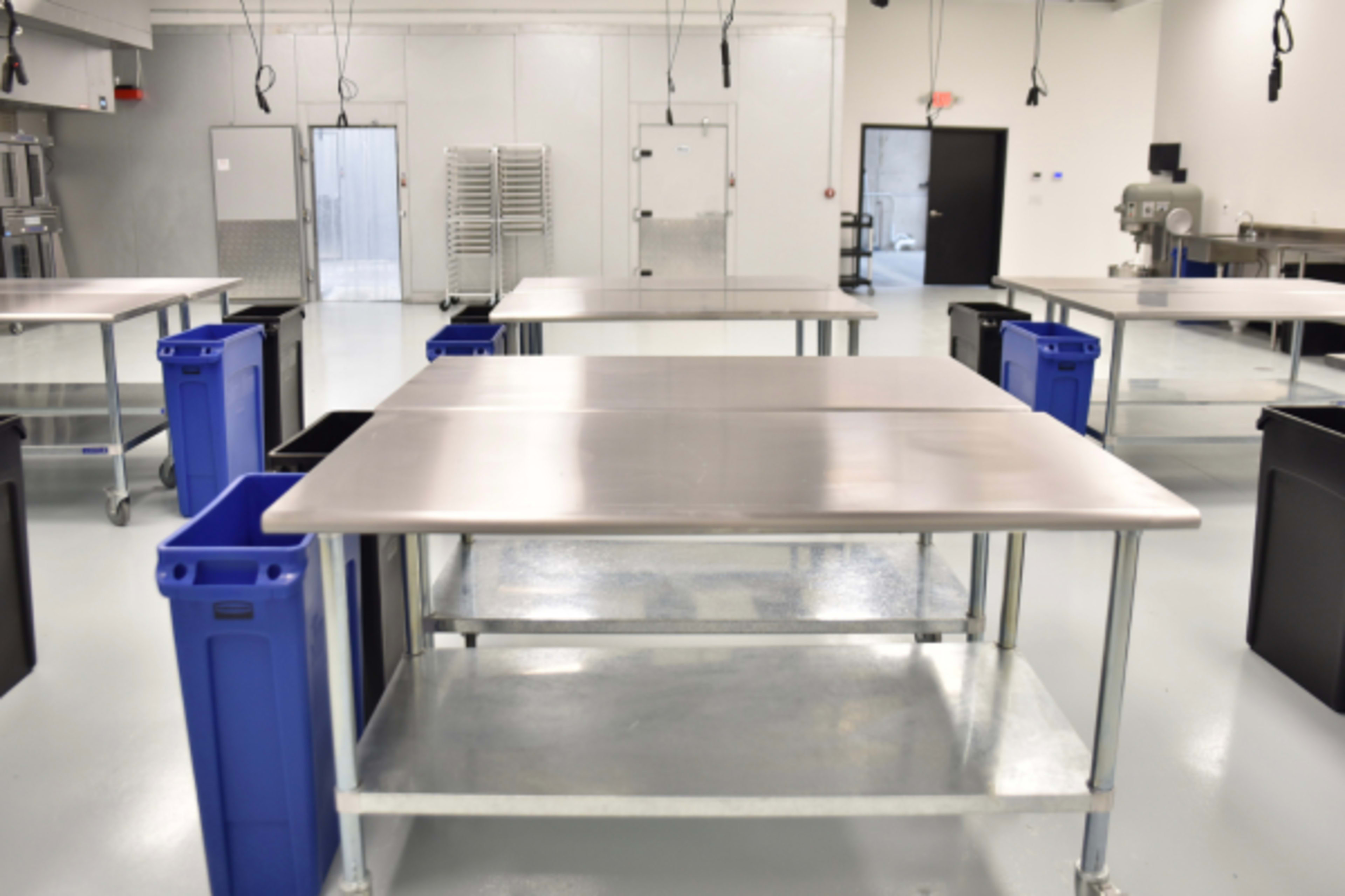 A clean commercial kitchen with stainless steel tables and blue recycling bins positioned on either side.