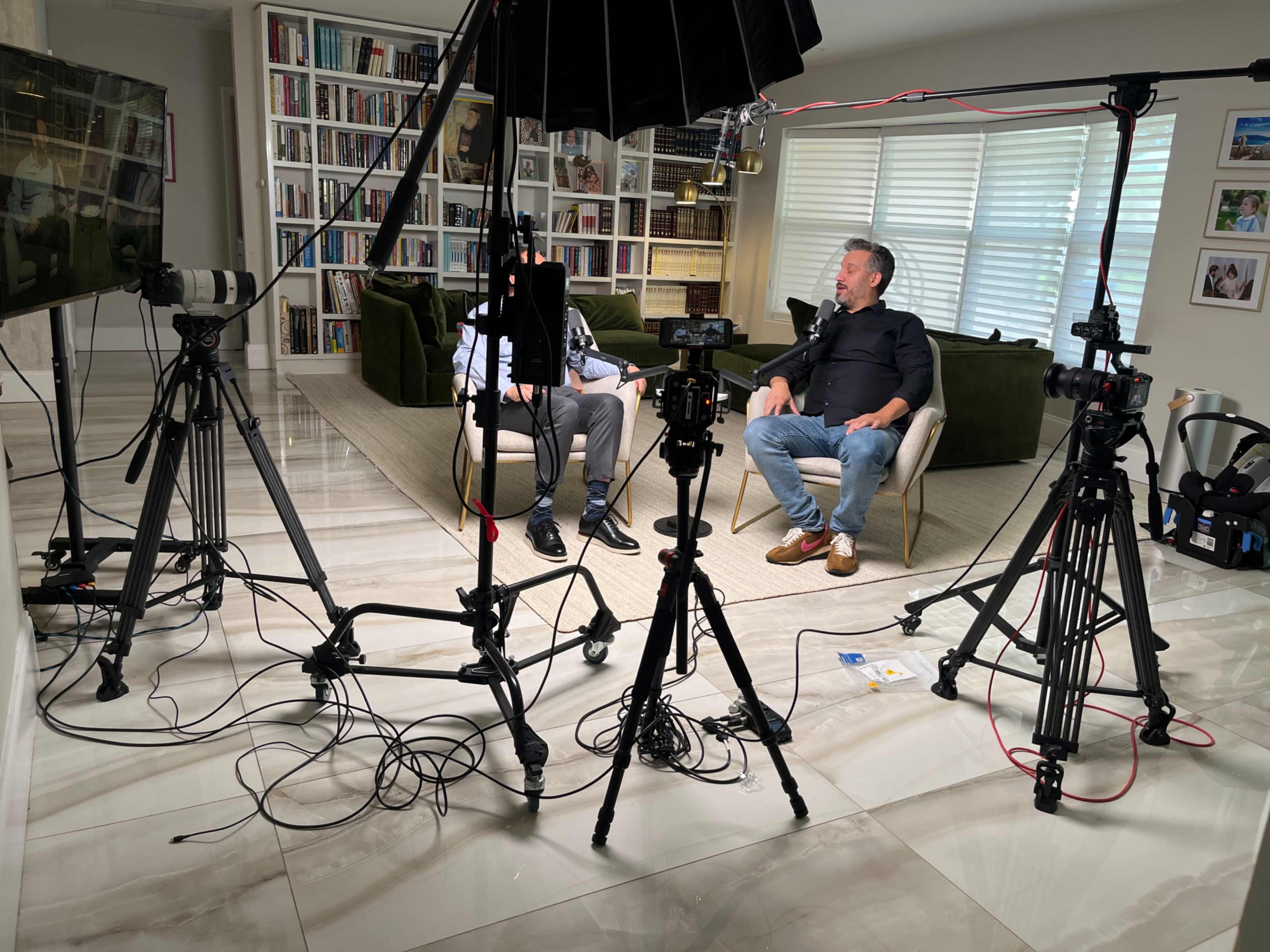 The image shows two men sitting on a couch in a well-lit room, surrounded by camera equipment and lighting setups for a video recording.