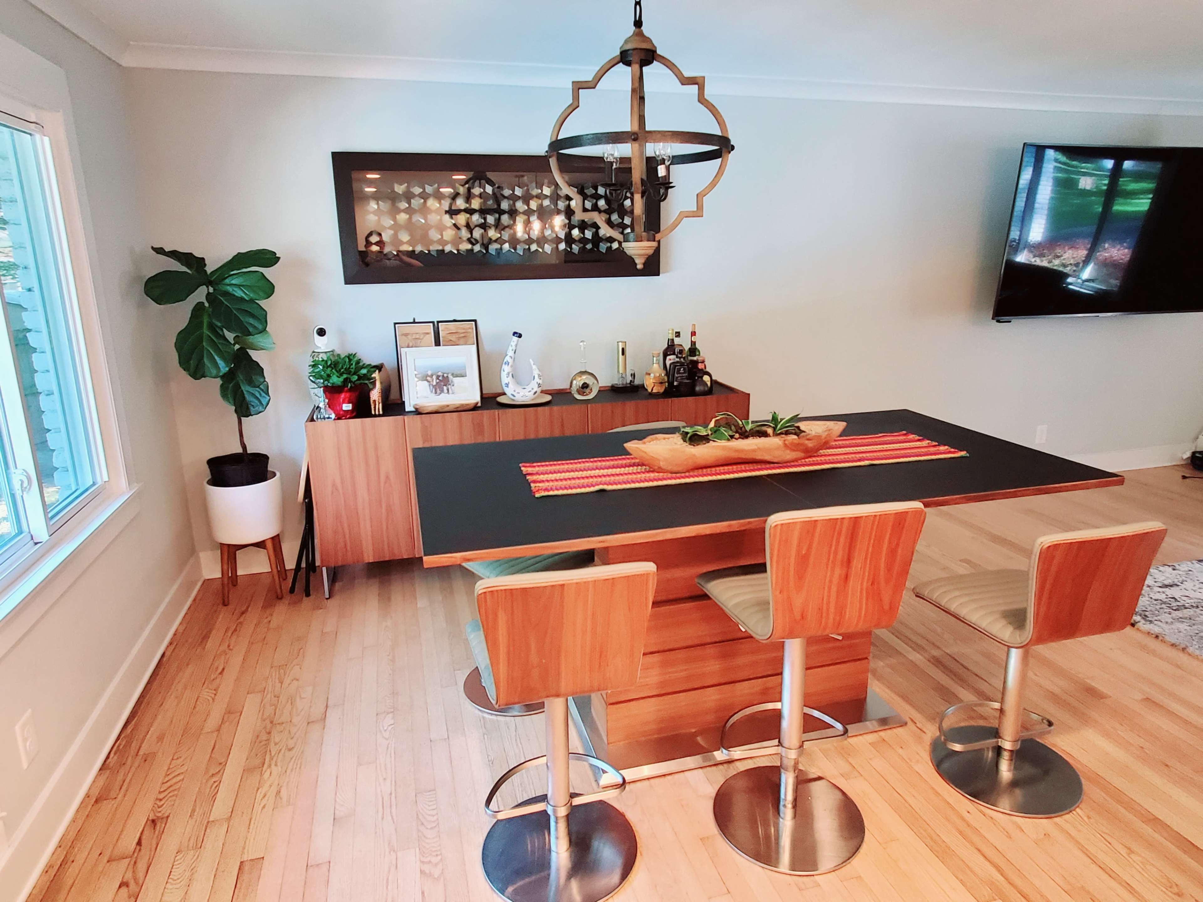 A modern dining area features a rectangular table with three stools, a wall-mounted display case, and a television in the background.