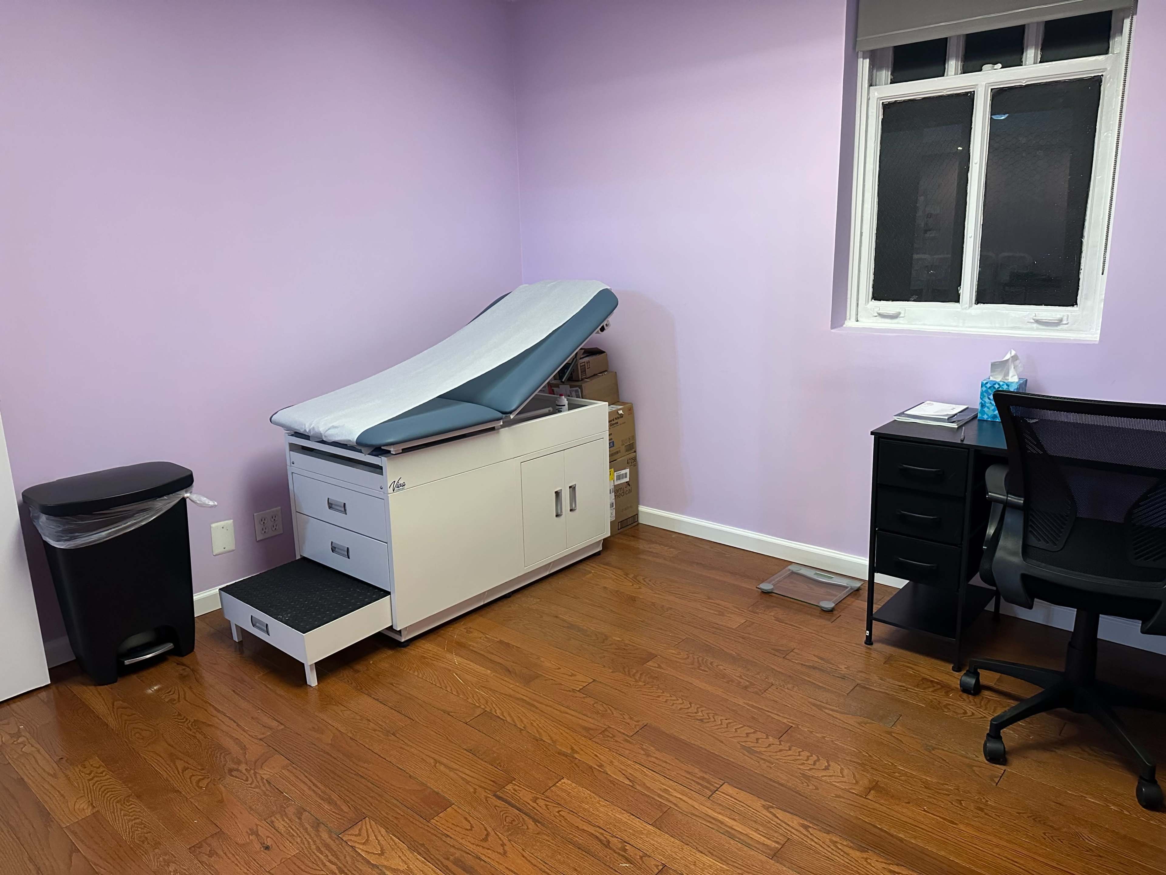 The image shows a medical examination room with a patient examination table, a waste bin, a black desk with a chair, and a window, all set against a purple wall.