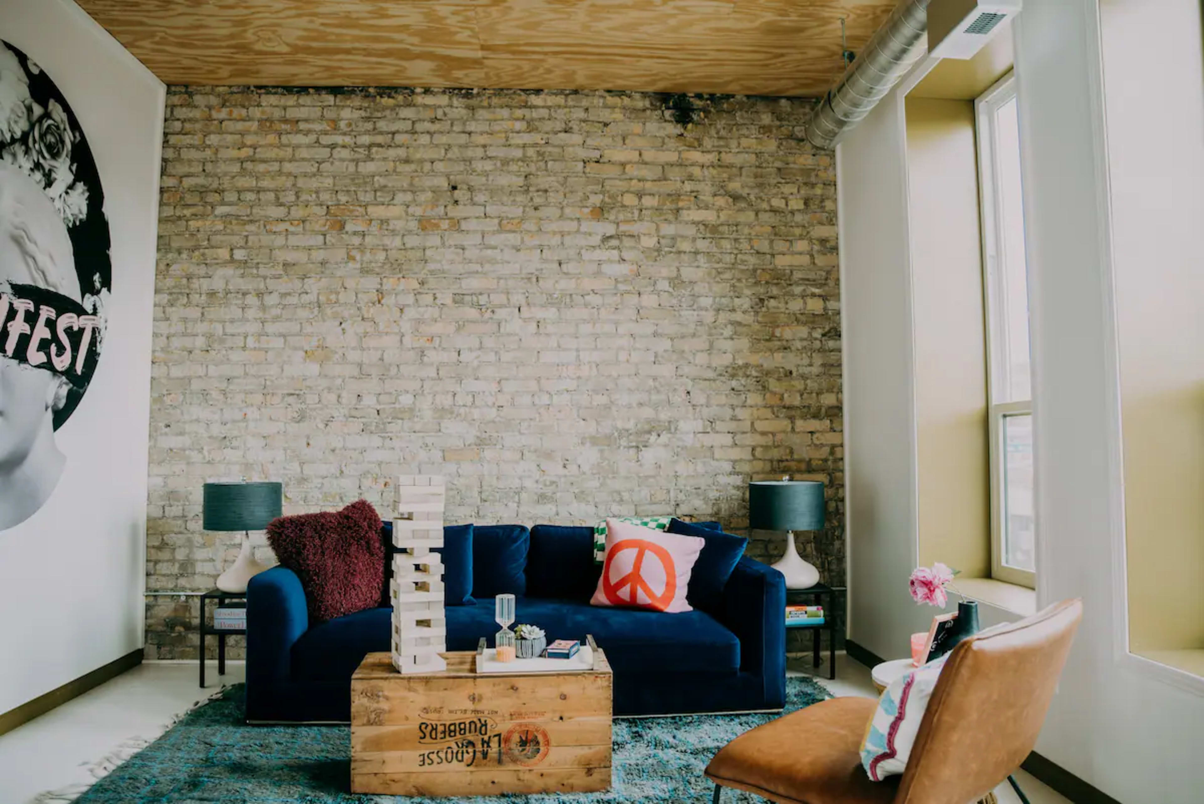 A cozy living room featuring a blue sofa, a wooden coffee table with games, and a decorative peace sign pillow, with exposed brick walls and large windows.