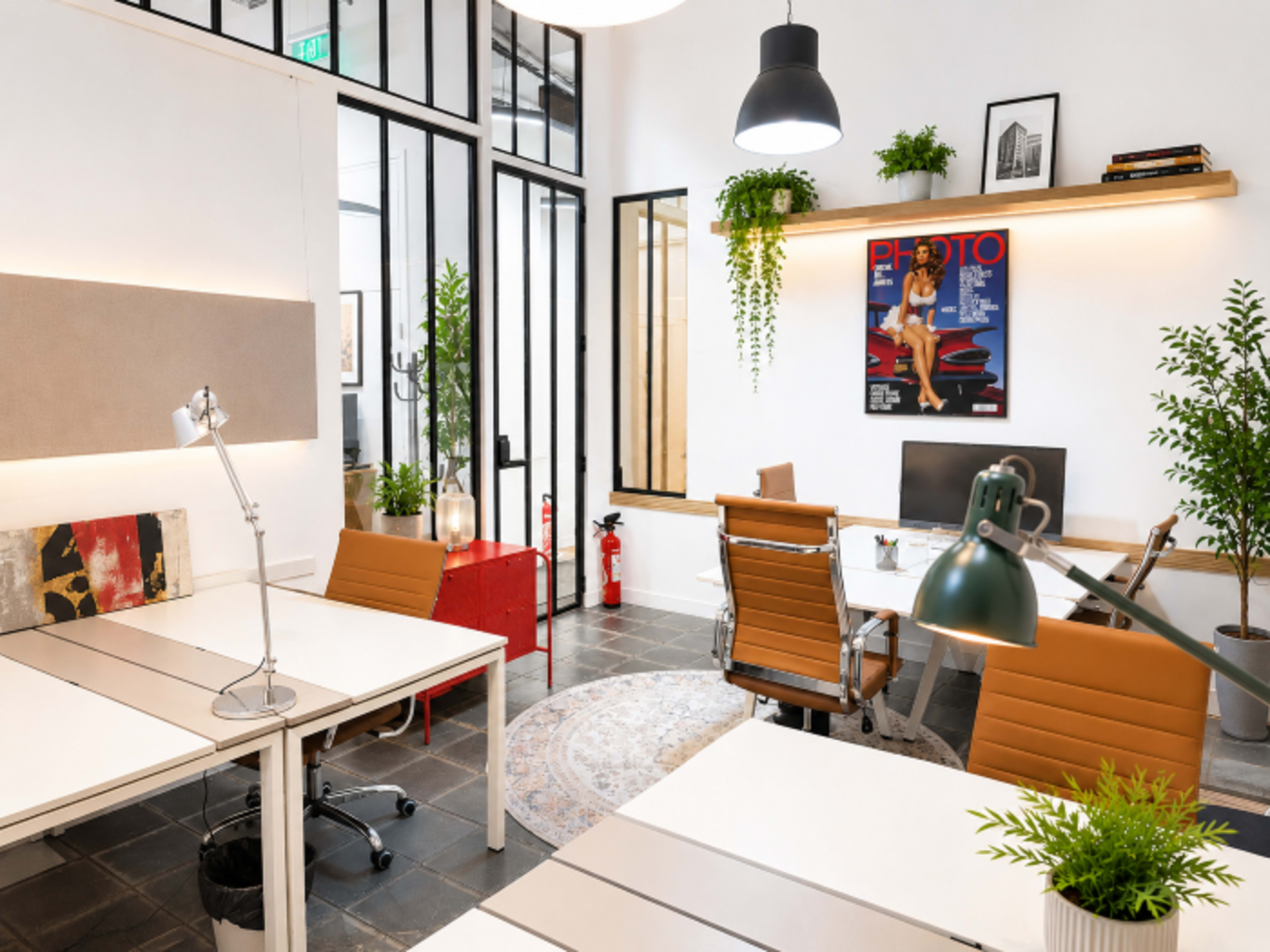The image shows a modern office space with desks, chairs, potted plants, and a computer, featuring large windows and artistic decor on the walls.