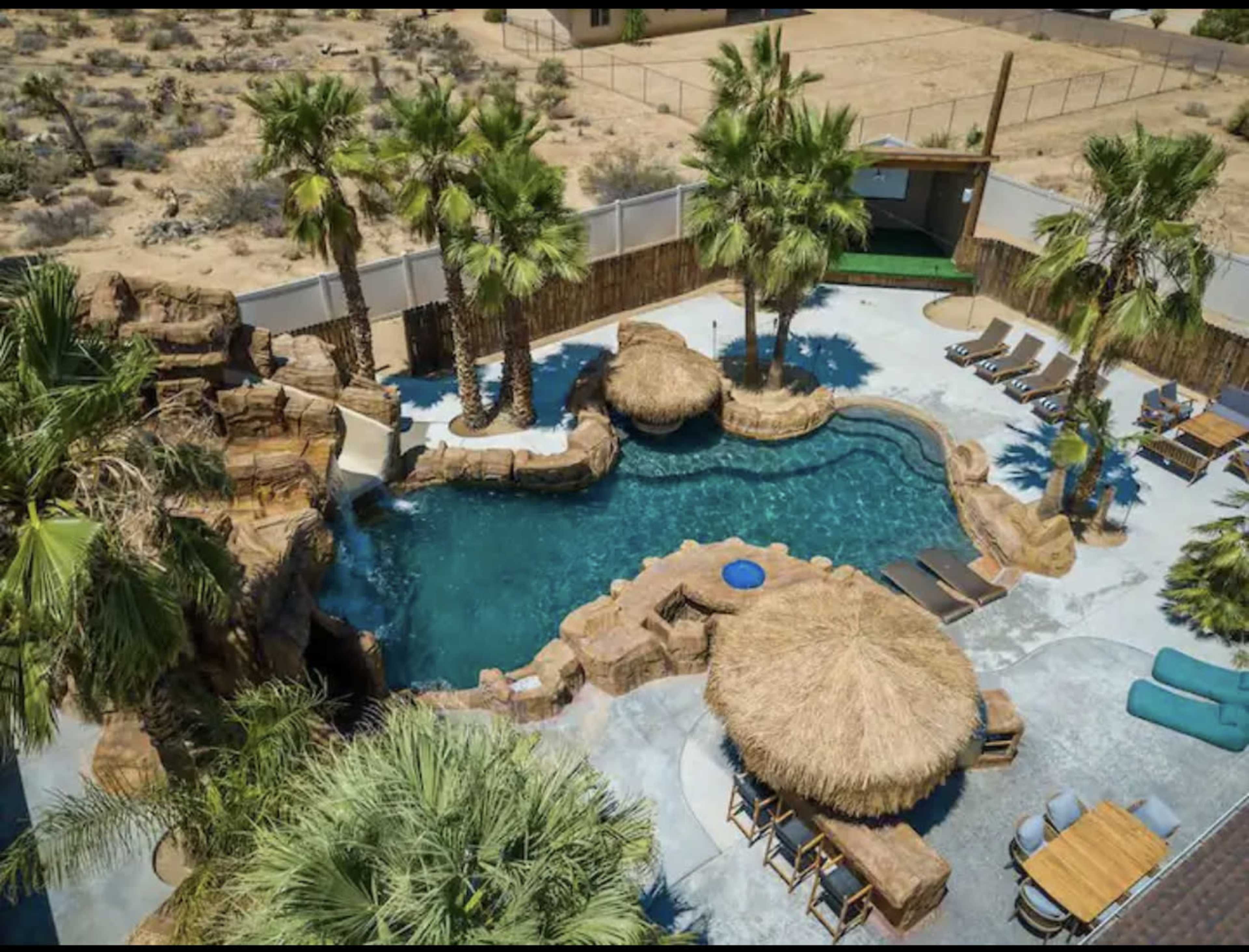 A landscaped pool area surrounded by palm trees, featuring rock formations, thatched umbrellas, and lounge chairs.
