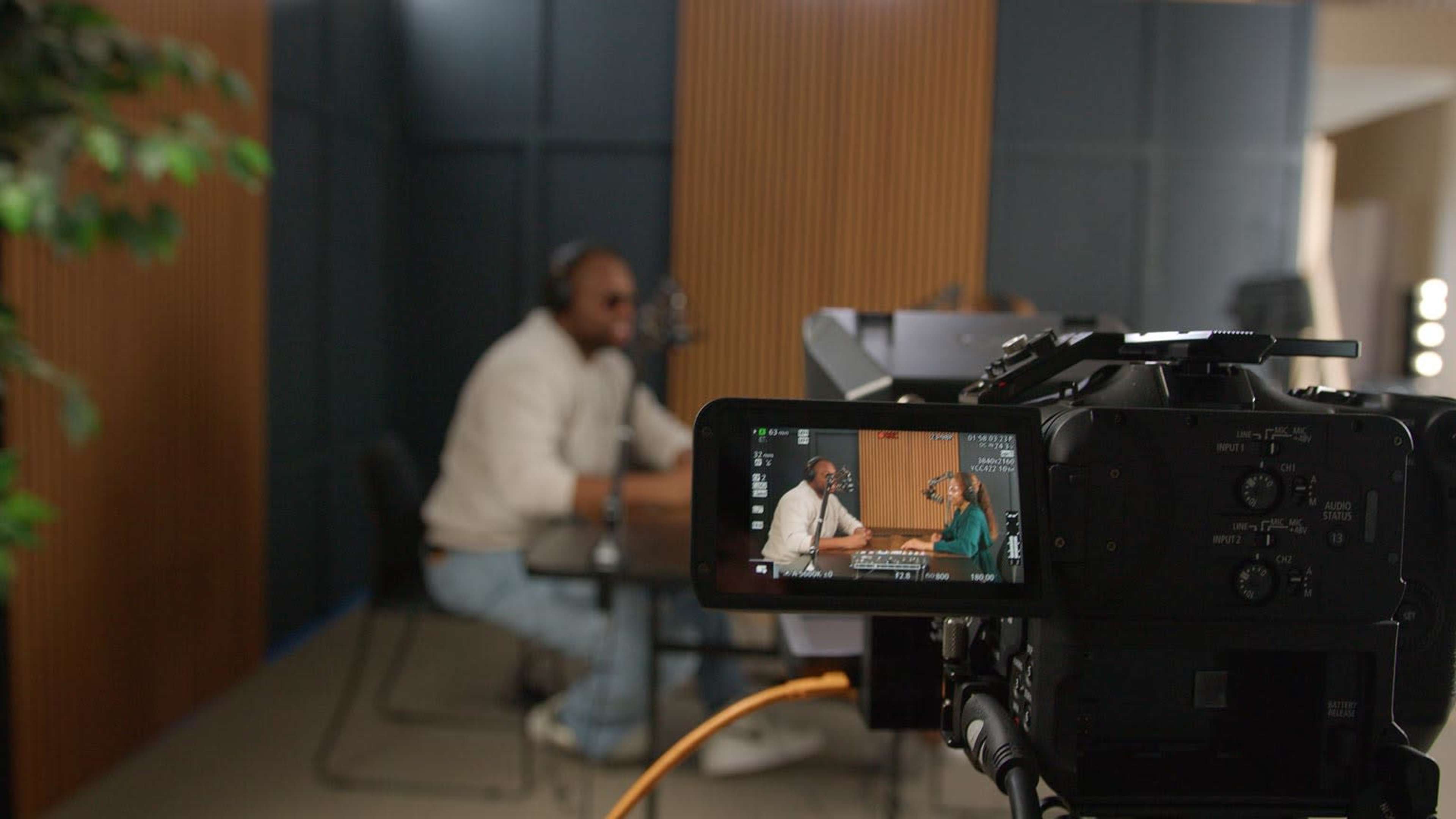 The image shows a camera focused on two individuals seated at a table, engaged in conversation in a well-lit studio environment.