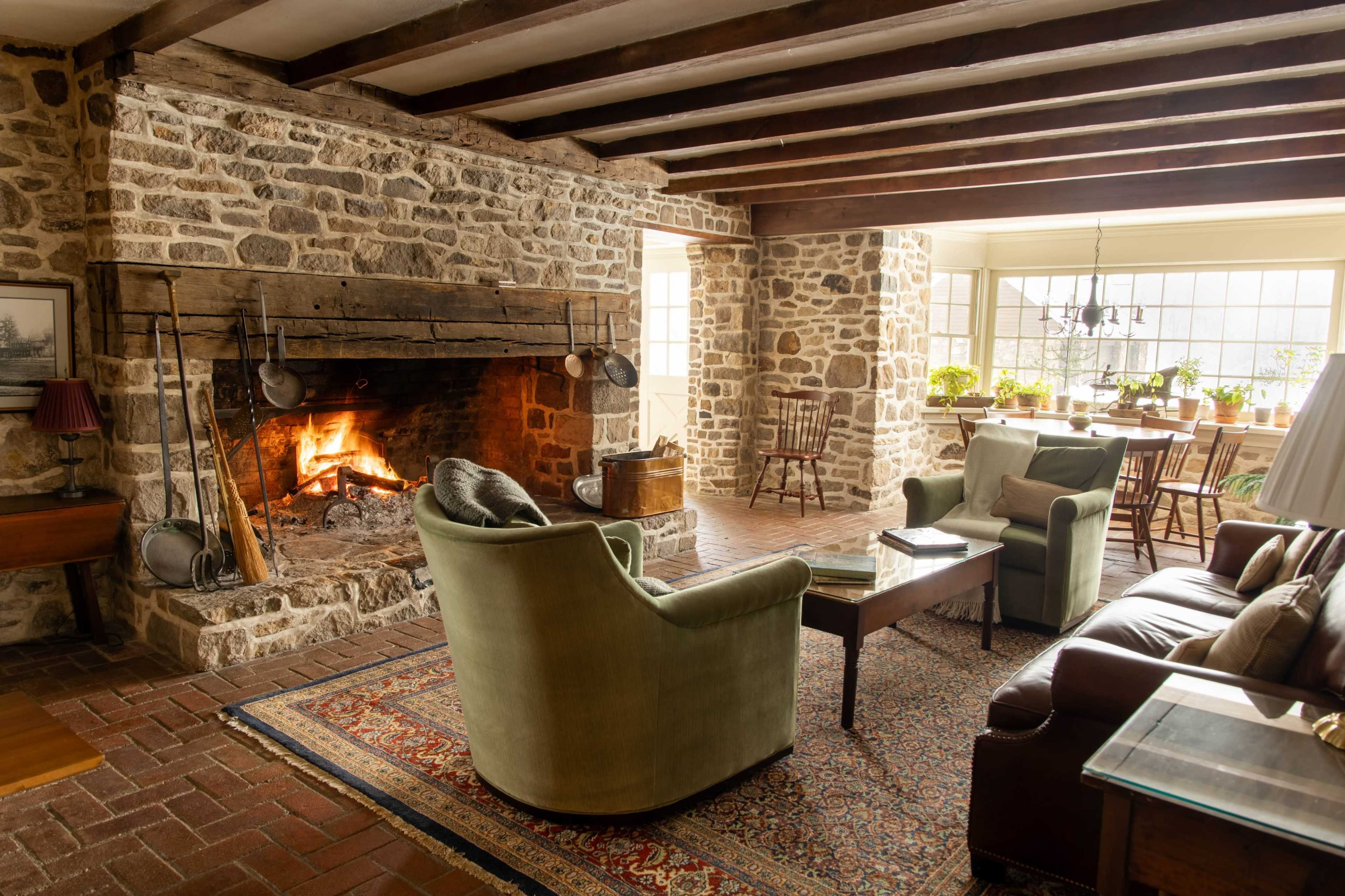 The image shows a cozy living room featuring a stone fireplace, two green chairs, a brown leather sofa, and large windows with plants.