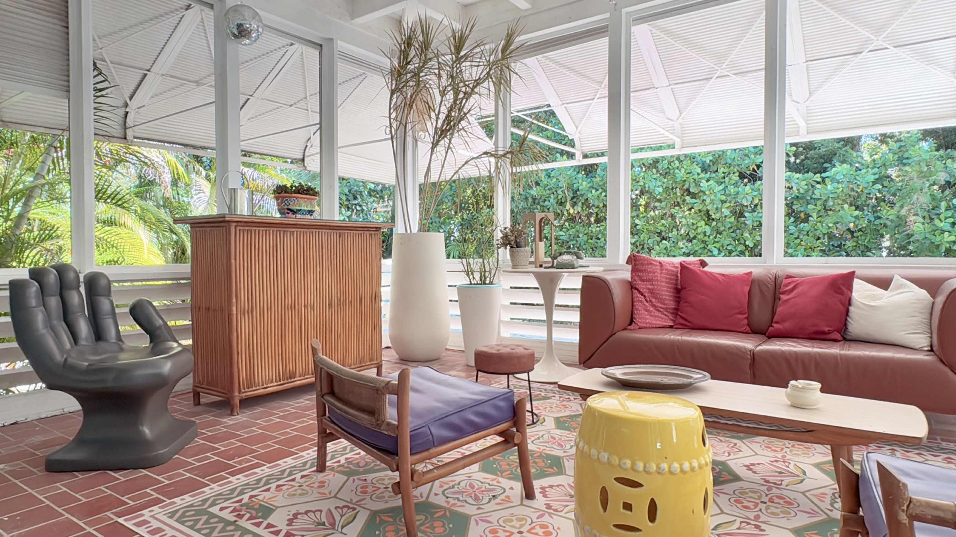 The image features a stylish outdoor living space with a mix of modern furniture, including a leather sofa, a contemporary chair, and decorative plants, surrounded by large windows that offer a view of greenery.