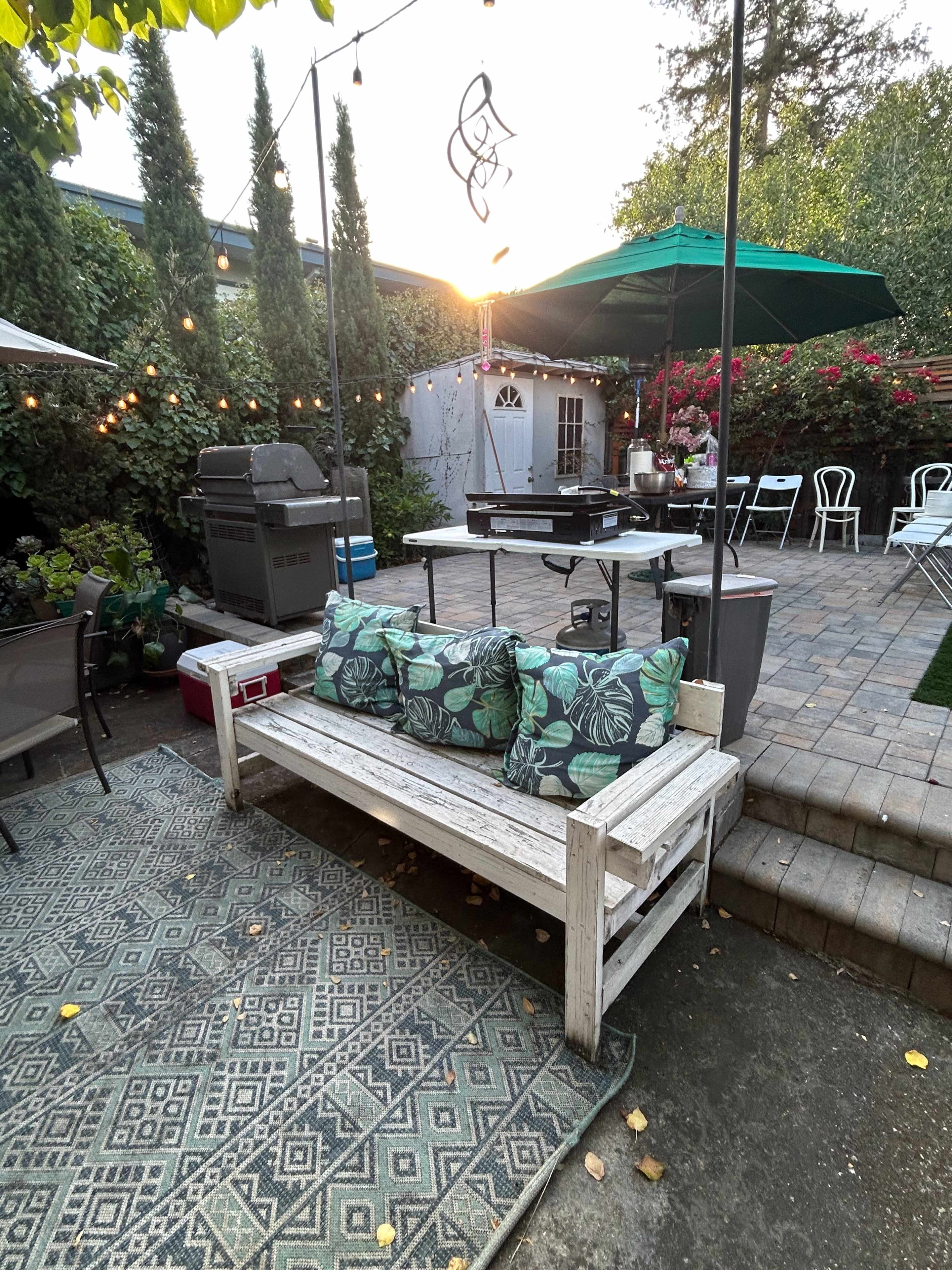 A wooden bench with decorative cushions is positioned on a patio surrounded by greenery, a barbecue grill, and tables set for outdoor dining.