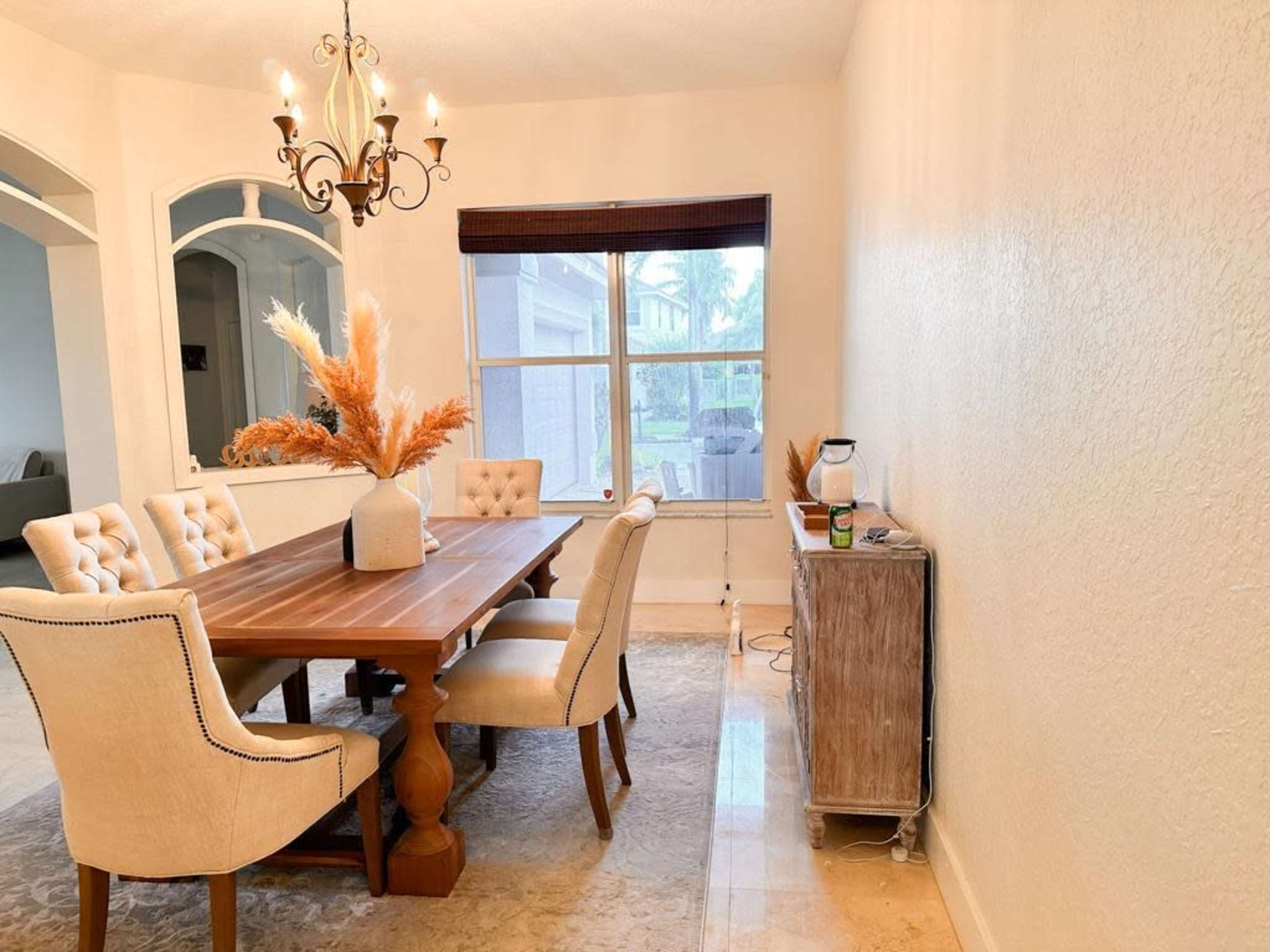 A dining room features a wooden table surrounded by beige upholstered chairs, with a chandelier overhead and decorative elements on the table.