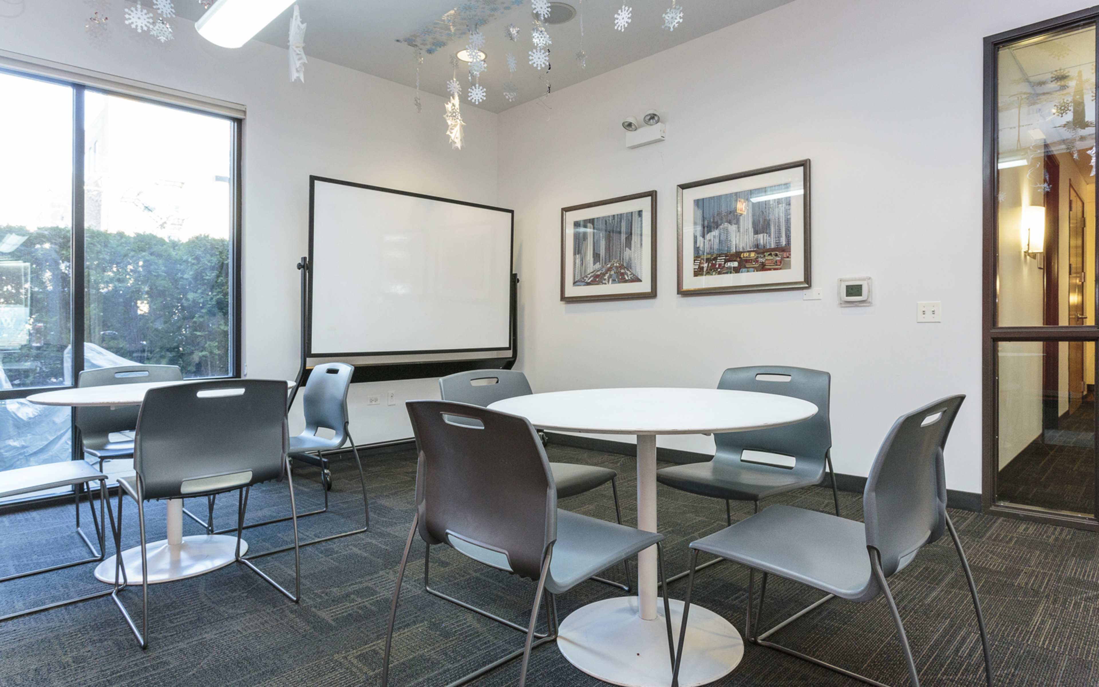 A small meeting room features a round table surrounded by six gray chairs and a large whiteboard on one wall, with framed pictures hanging beside it.