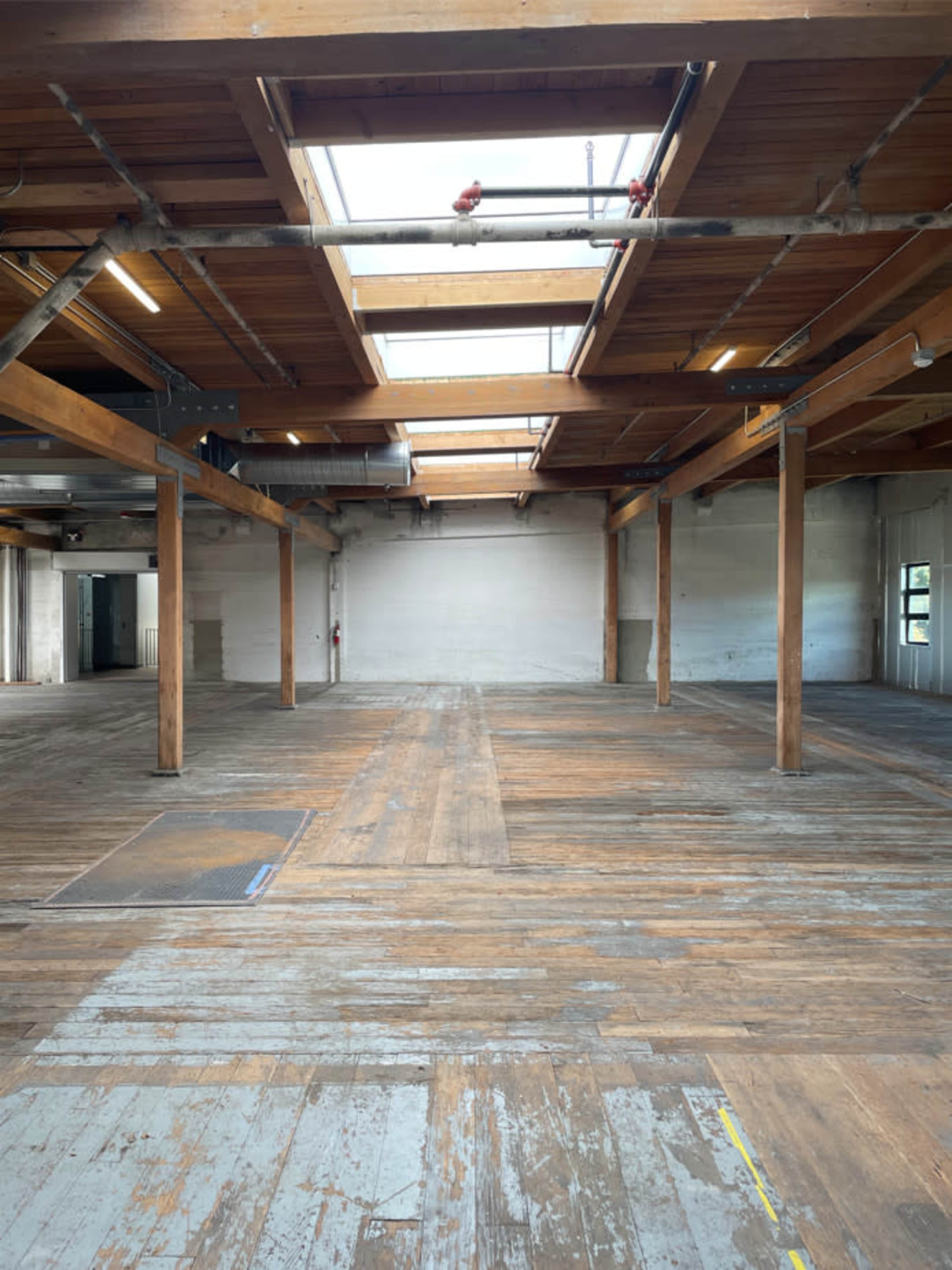 The image shows a spacious, empty room with wooden beams and a wooden floor, featuring skylights and several support columns.