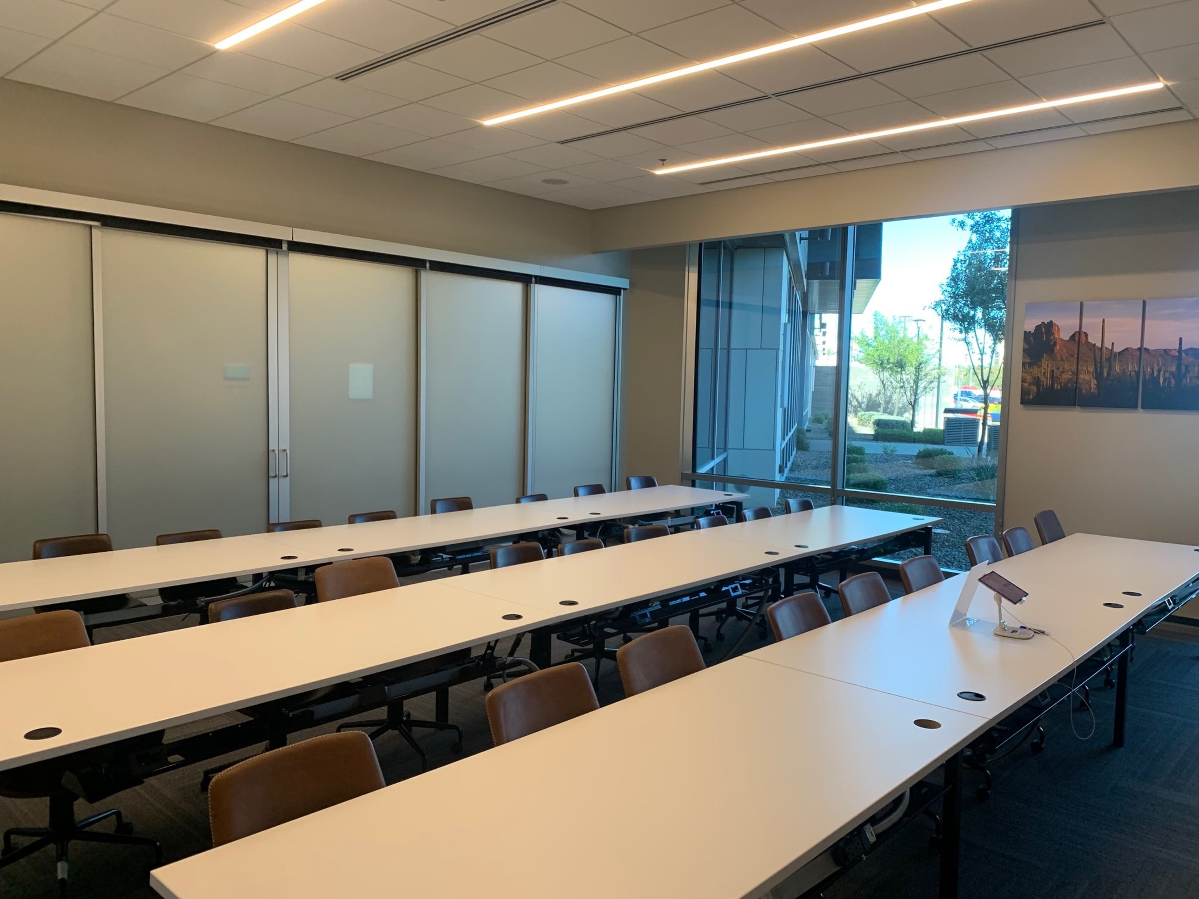 The image shows a conference room with multiple long tables arranged in a U-shape, featuring brown chairs and large windows overlooking an outdoor area.