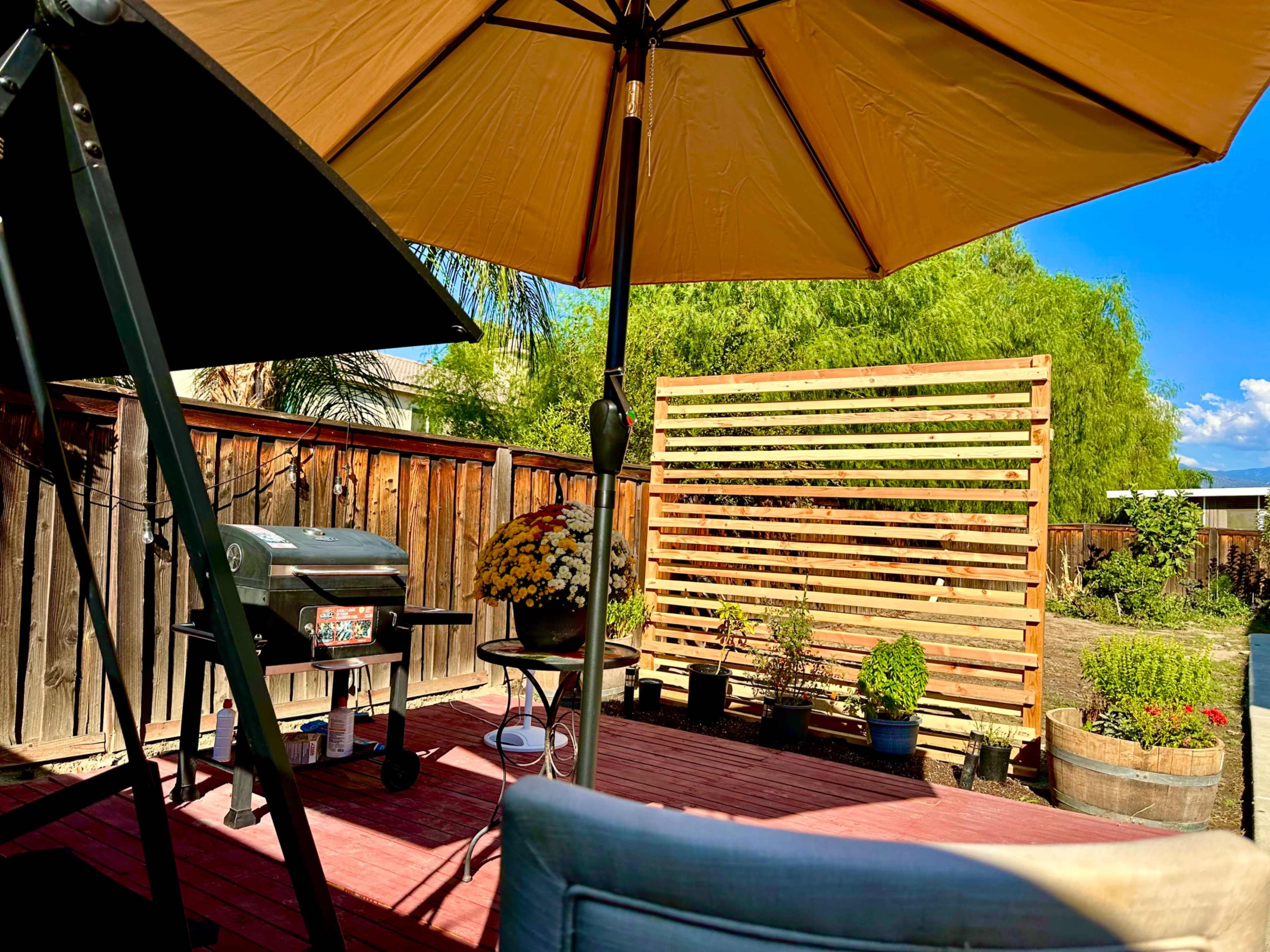 The image shows a patio area with a grill, a table with decorative plants, and an umbrella providing shade.