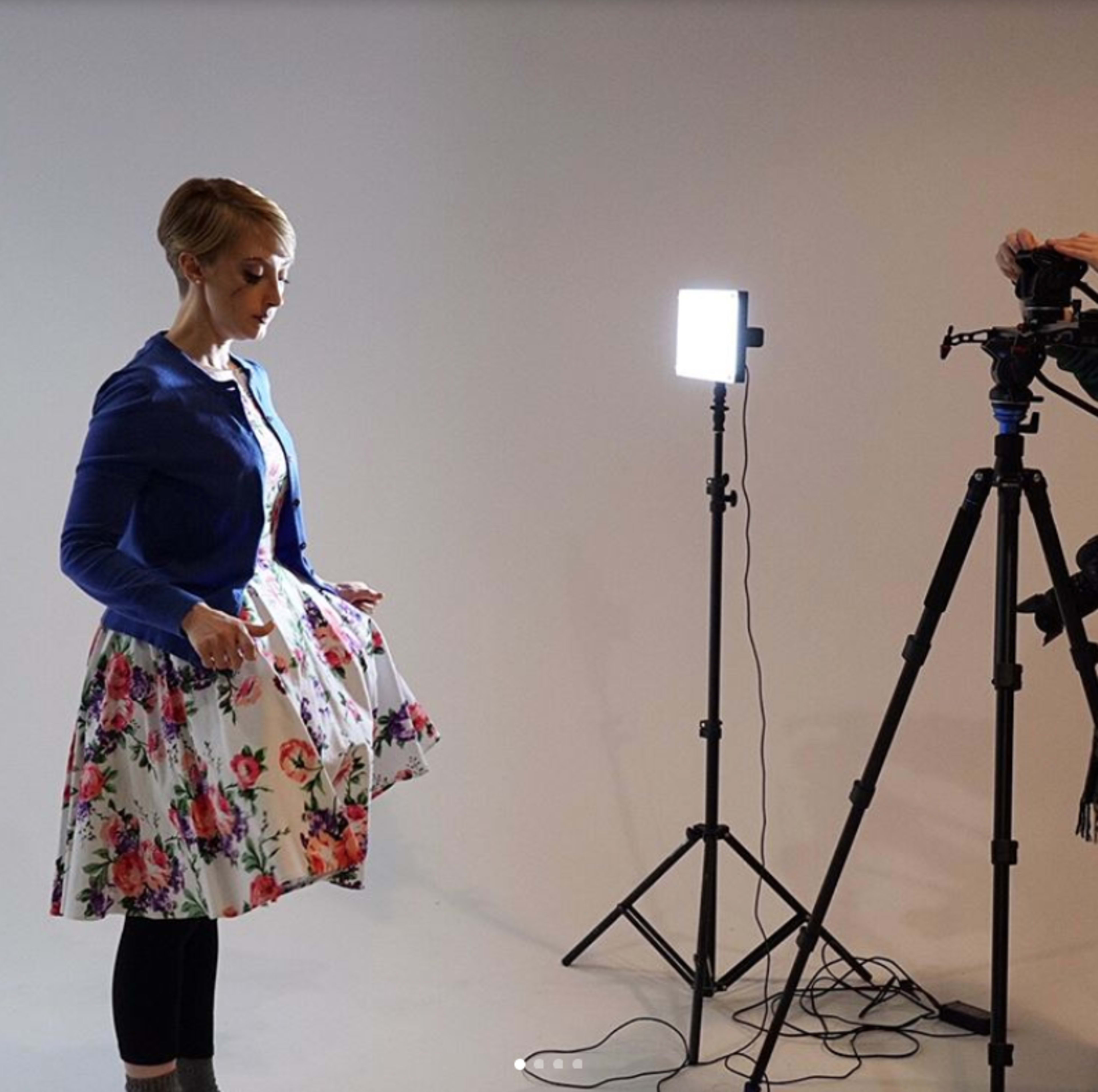 A woman in a floral dress adjusts her outfit in front of a camera set on a tripod, with a bright light illuminating the scene.