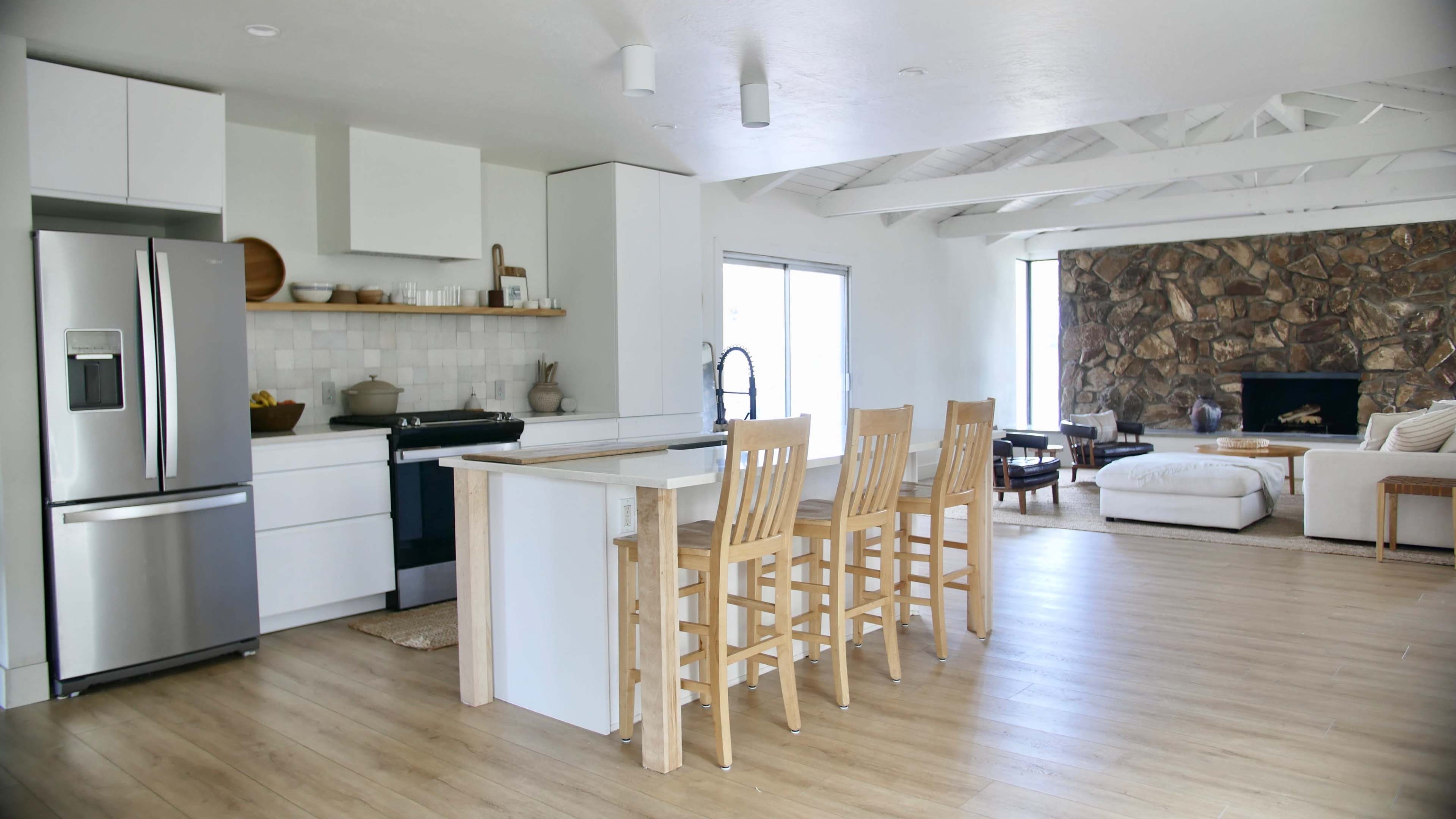 A modern kitchen with a central island and wooden bar stools, open to a living area featuring a stone fireplace and large windows.