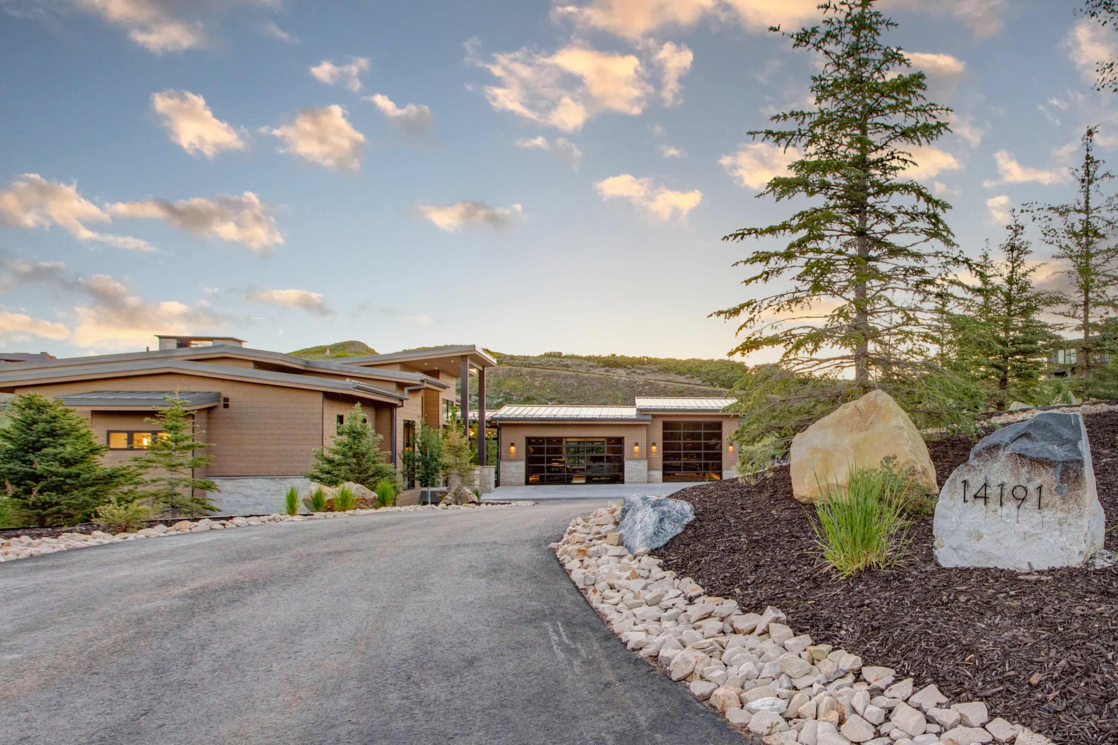 A modern house with a sleek design situated at the end of a driveway, surrounded by landscaped greenery and decorative rocks.