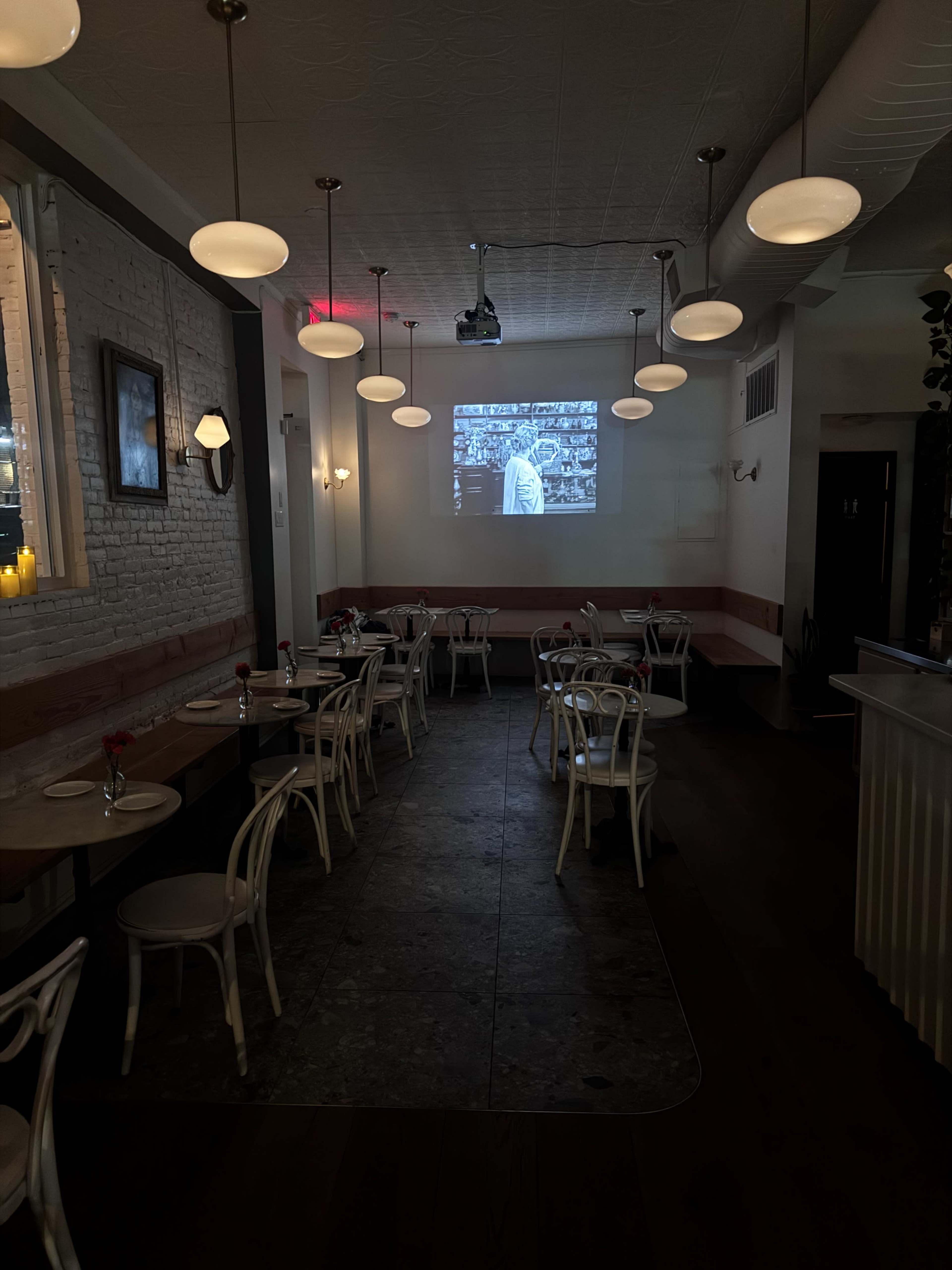 The image shows a dimly lit restaurant with white walls, several tables and chairs, and a projection screen displaying a black-and-white image on one wall.