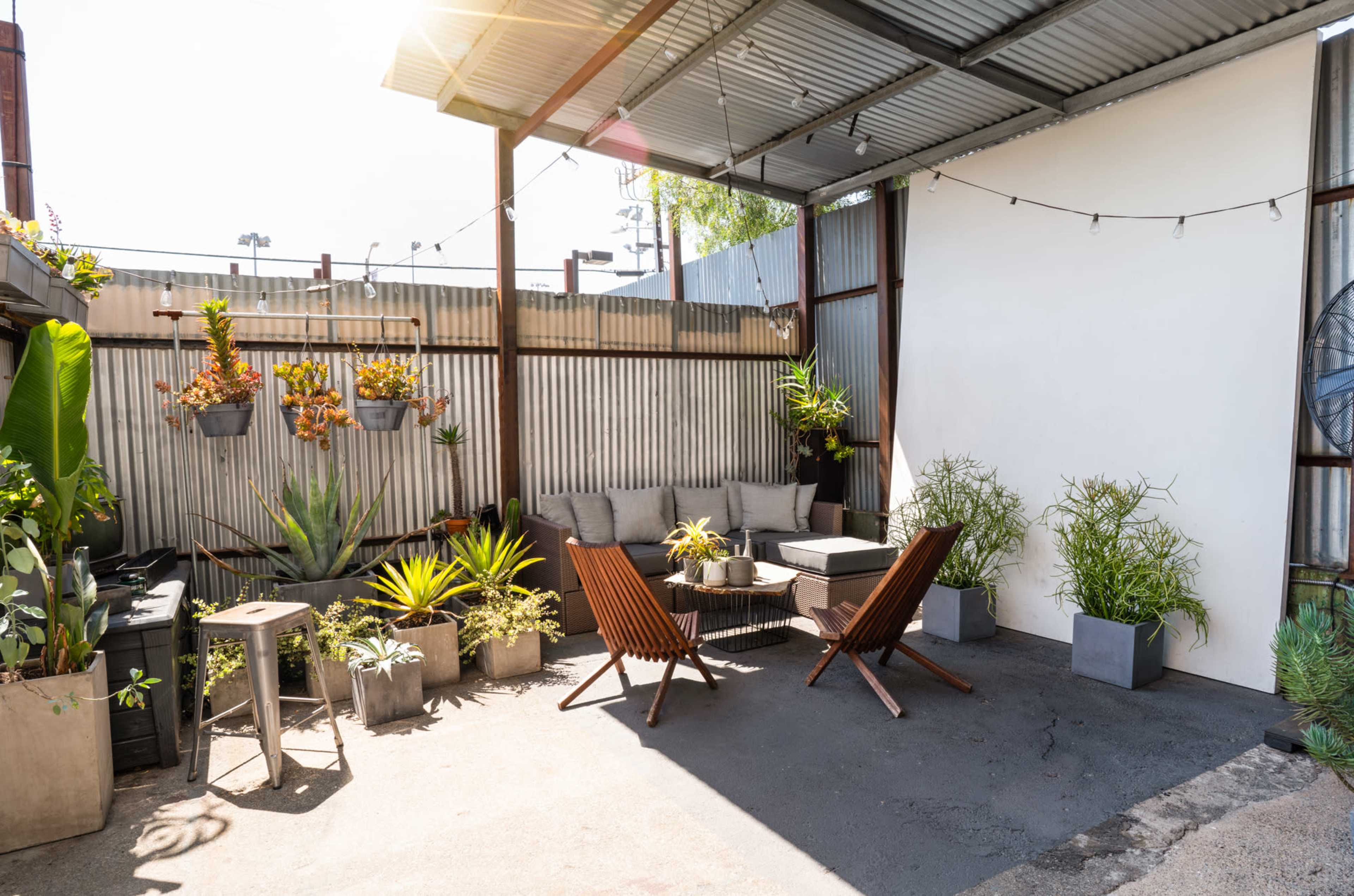 The image shows a small, outdoor seating area with two wooden lounge chairs, a sectional sofa, potted plants, and string lights under a metal canopy.