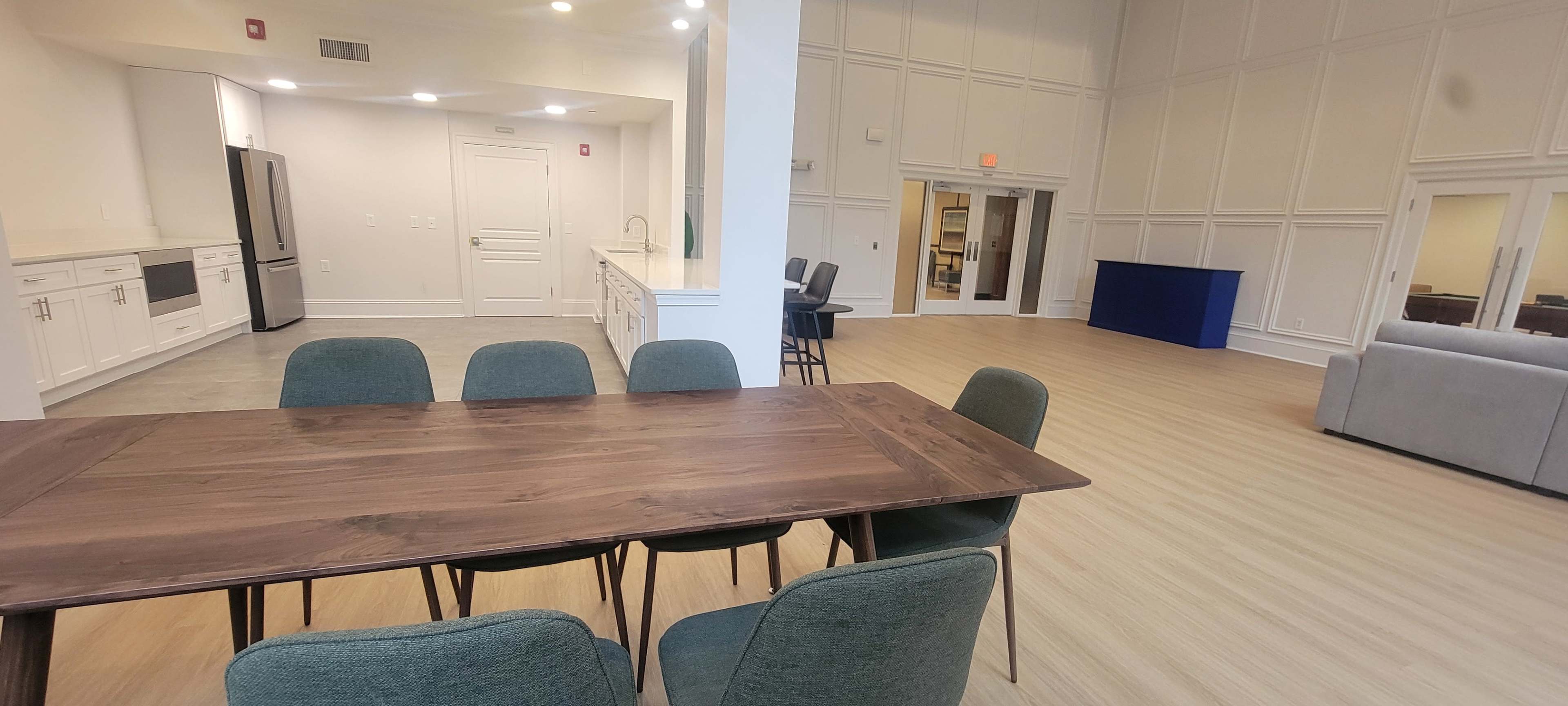 The image shows a modern communal room with a wooden dining table and chairs, a kitchen area with appliances on one side, and an open space featuring a seating area and a blue partitioned section.